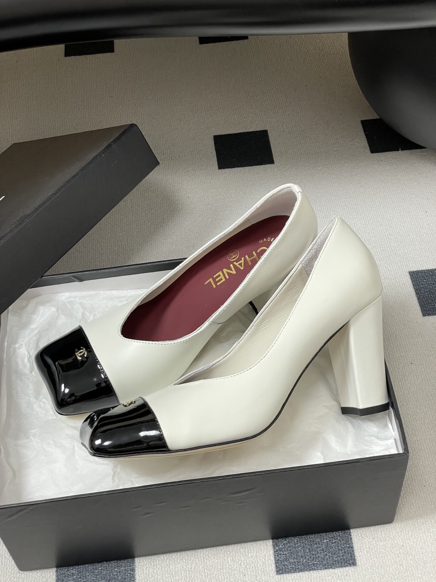 A pair of Chanel two-tone block heel pumps in white leather with black patent square toes and gold CC logos on a black table. - Hình 4