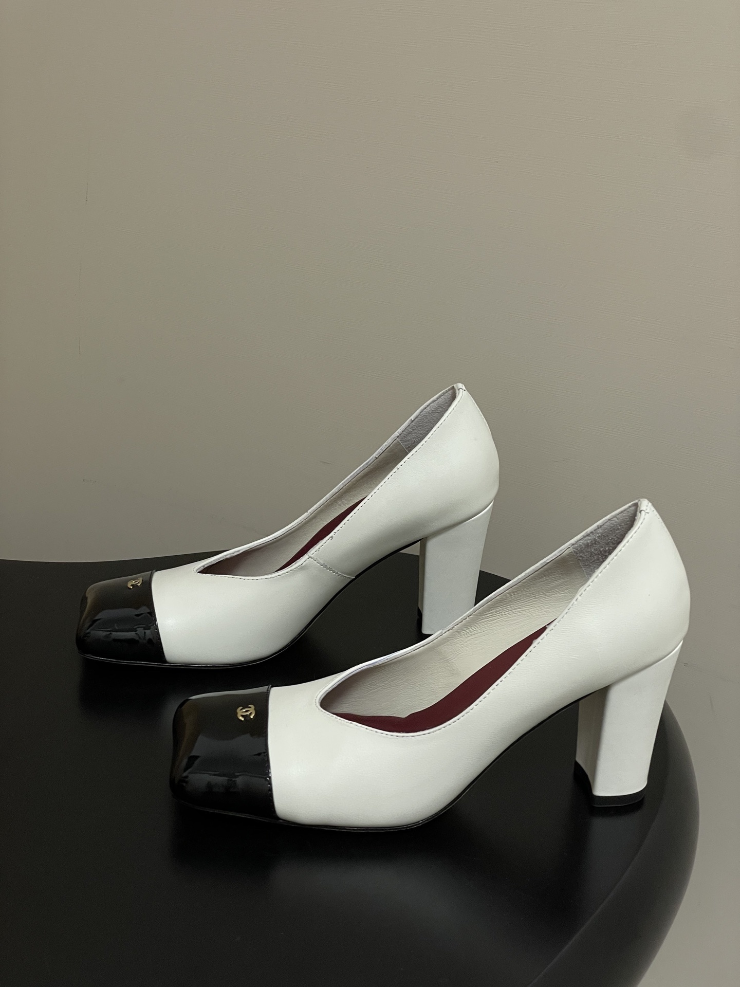 A pair of Chanel two-tone block heel pumps in white leather with black patent square toes and gold CC logos on a black table. - Hình 3