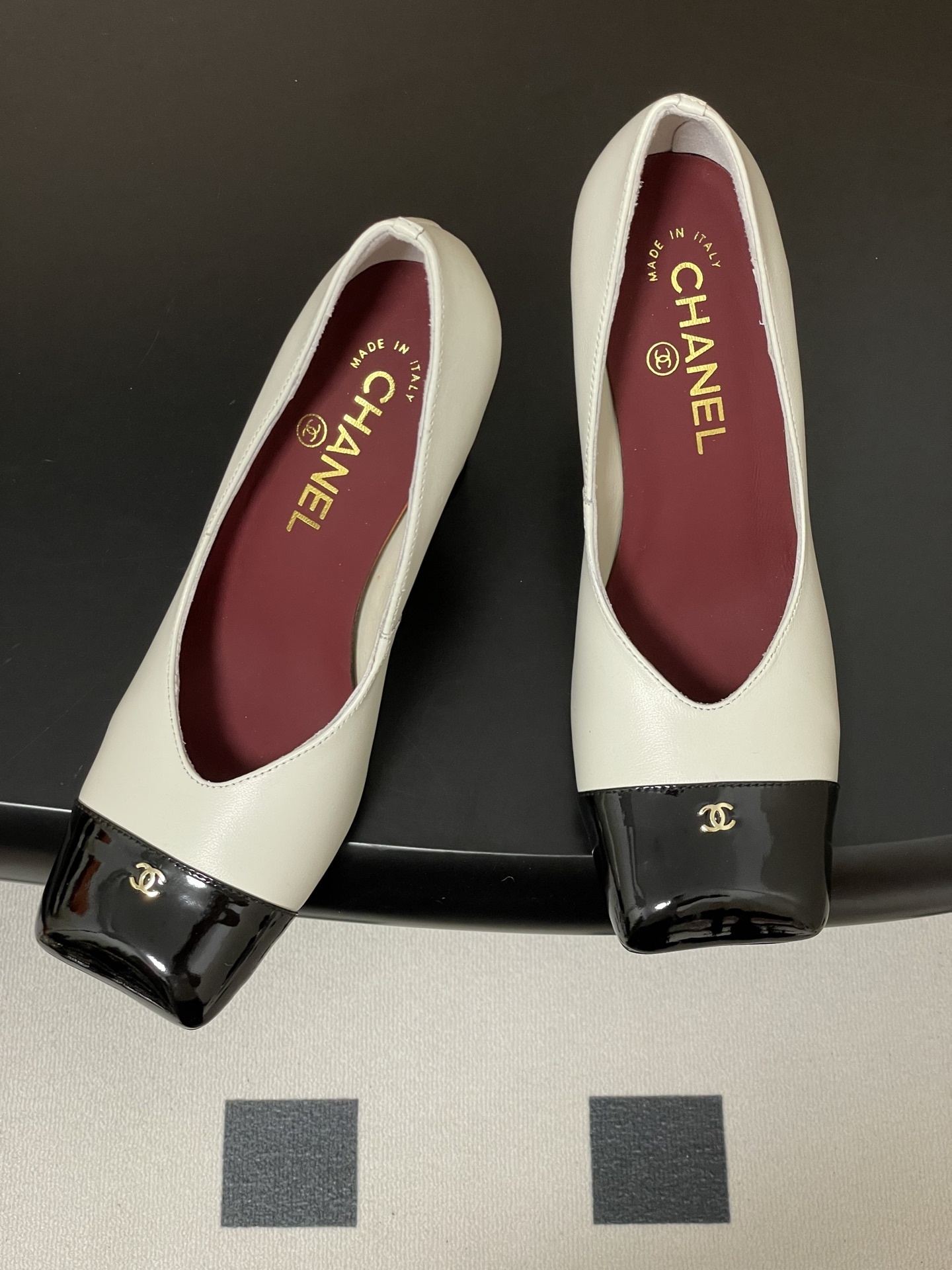 A pair of Chanel two-tone block heel pumps in white leather with black patent square toes and gold CC logos on a black table. - Hình 2