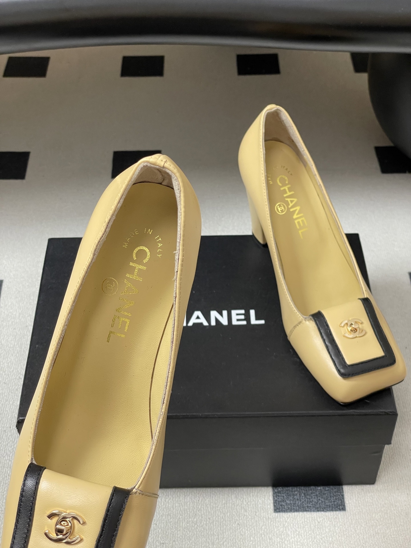 A pair of luxury Chanel beige leather block heel pumps with black trim and gold CC logo detailing on a black surface. - Hình 7