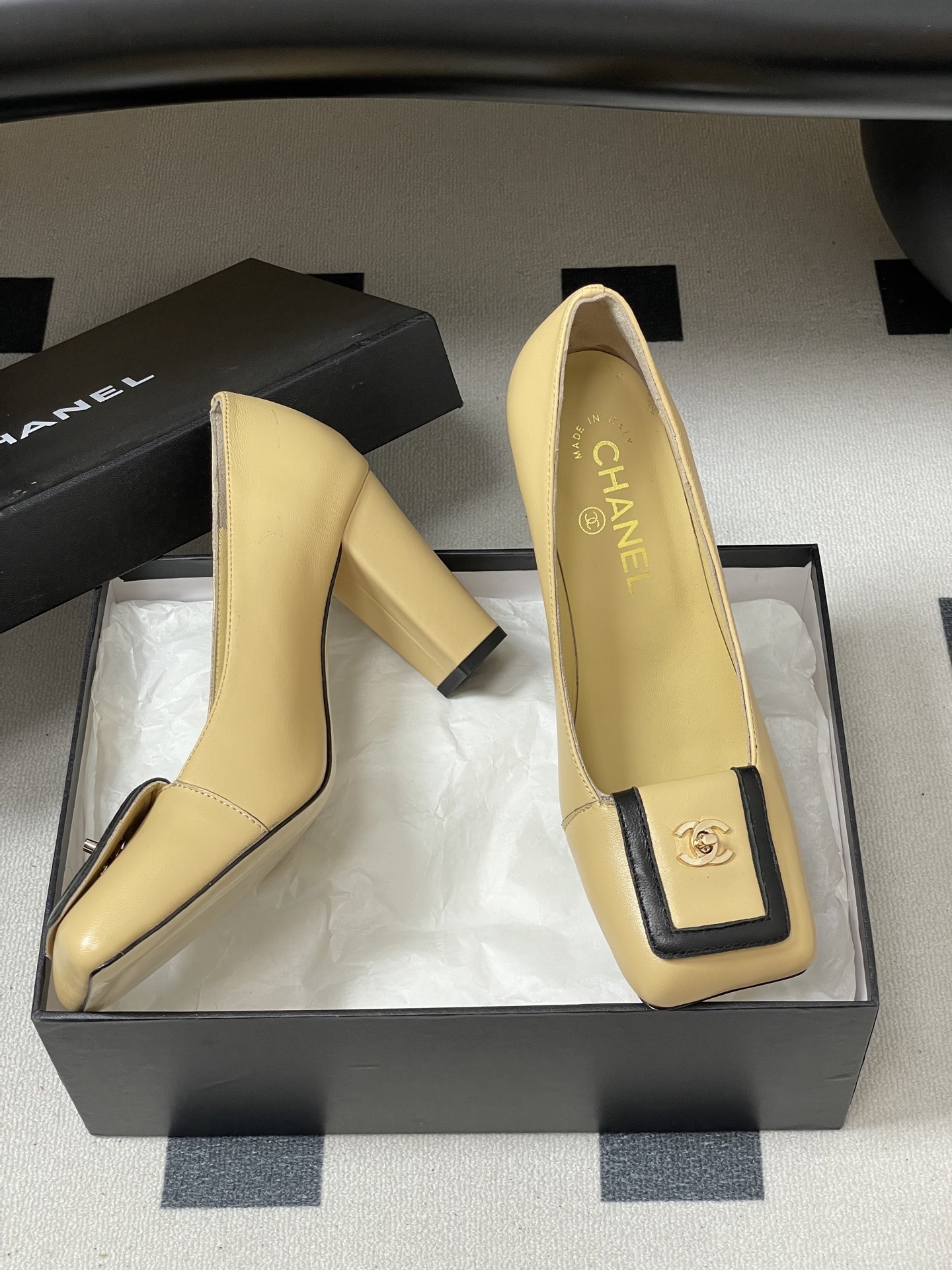 A pair of luxury Chanel beige leather block heel pumps with black trim and gold CC logo detailing on a black surface. - Hình 6