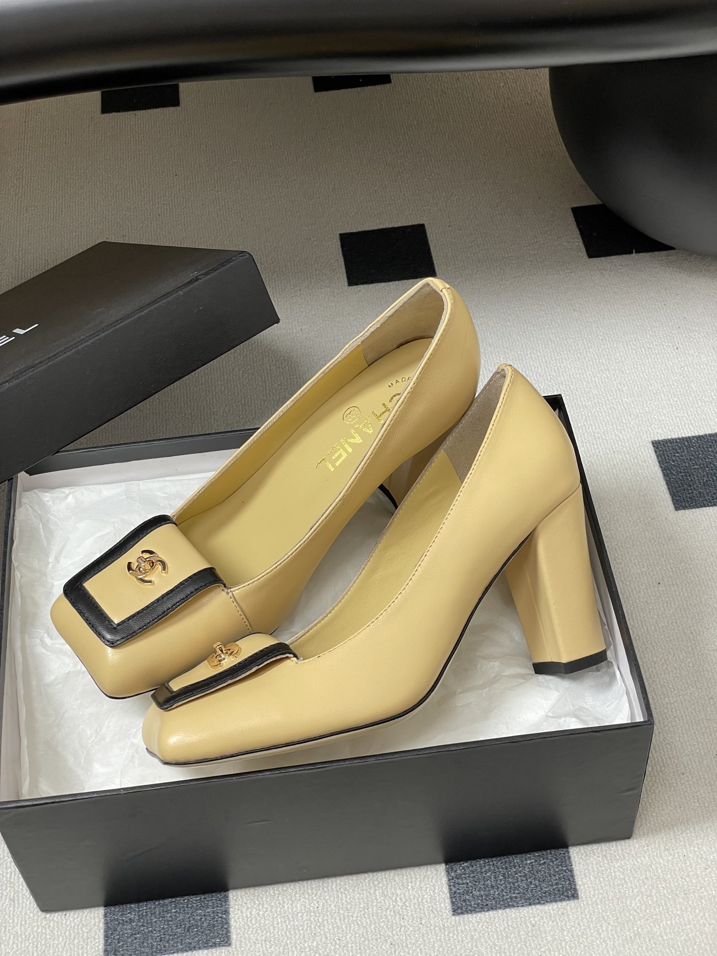 A pair of luxury Chanel beige leather block heel pumps with black trim and gold CC logo detailing on a black surface. - Hình 4