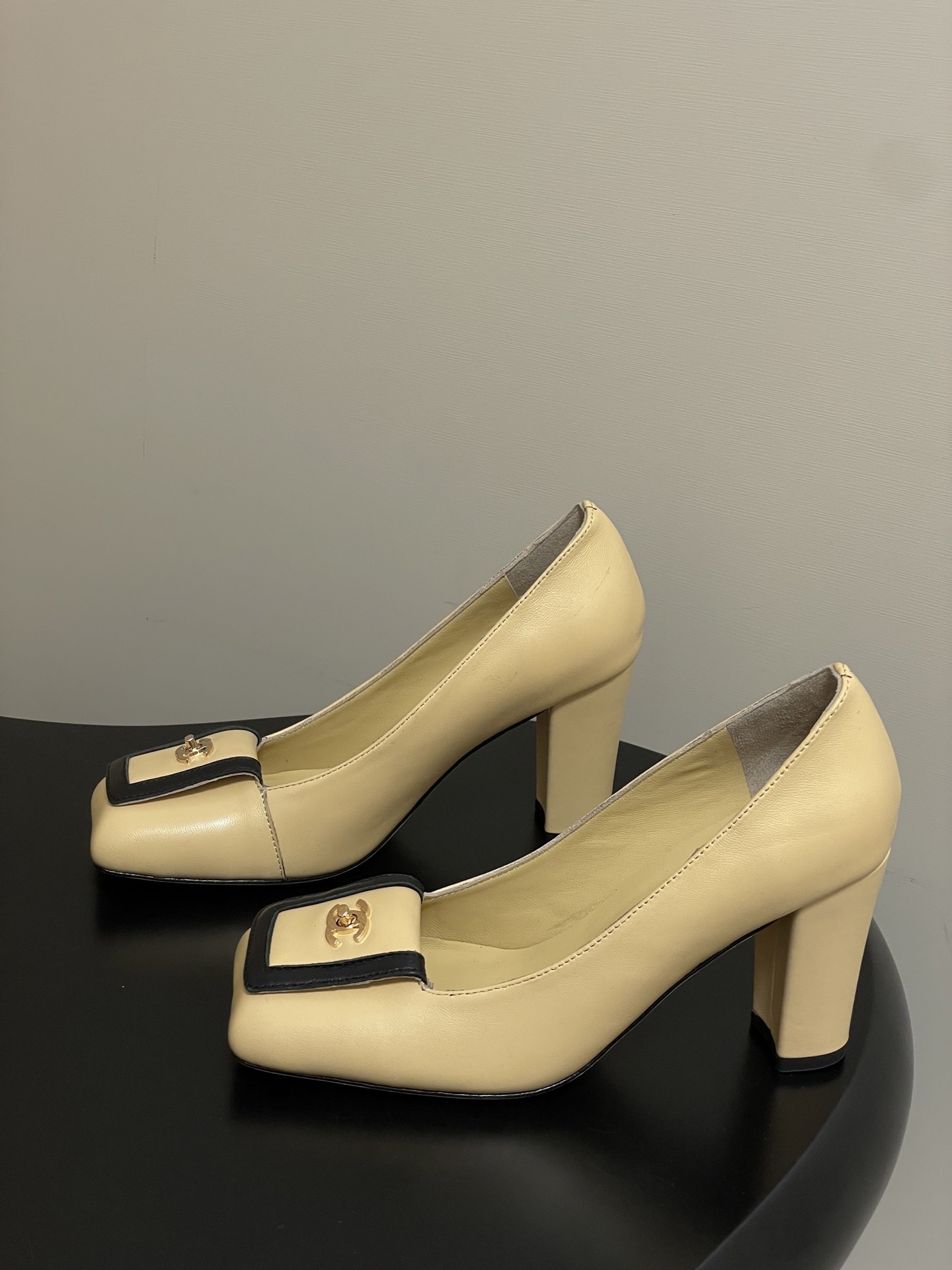 A pair of luxury Chanel beige leather block heel pumps with black trim and gold CC logo detailing on a black surface. - Hình 3