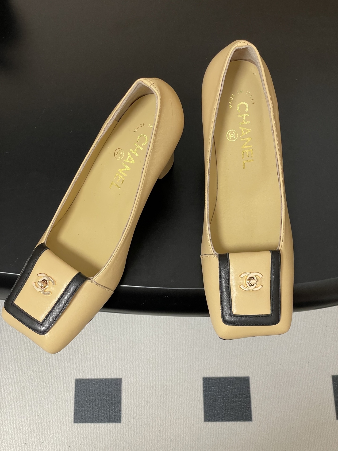 A pair of luxury Chanel beige leather block heel pumps with black trim and gold CC logo detailing on a black surface. - Hình 2