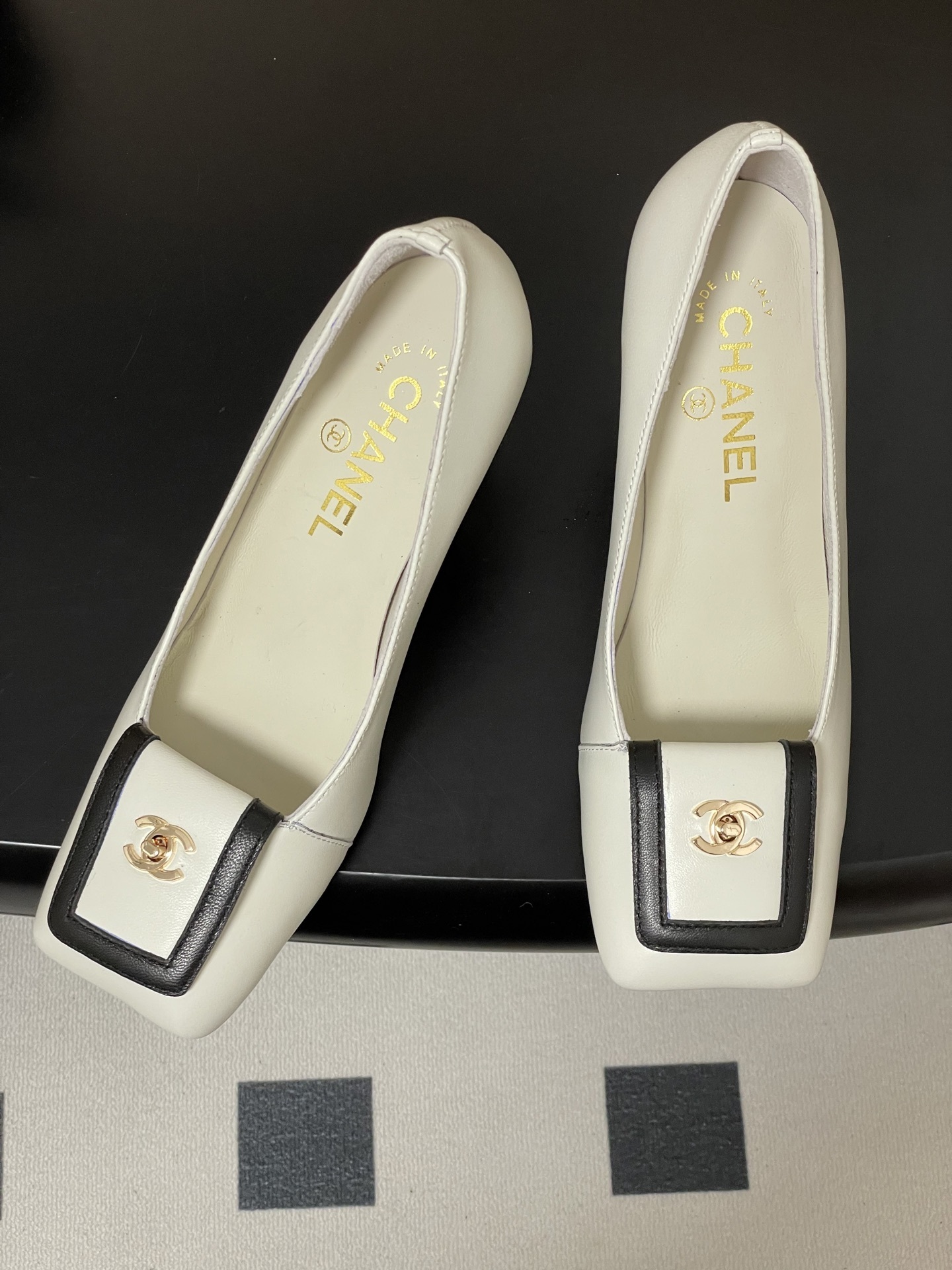 Pair of Chanel white leather square toe pumps with black trim and gold CC logo on a black surface. - Hình 2