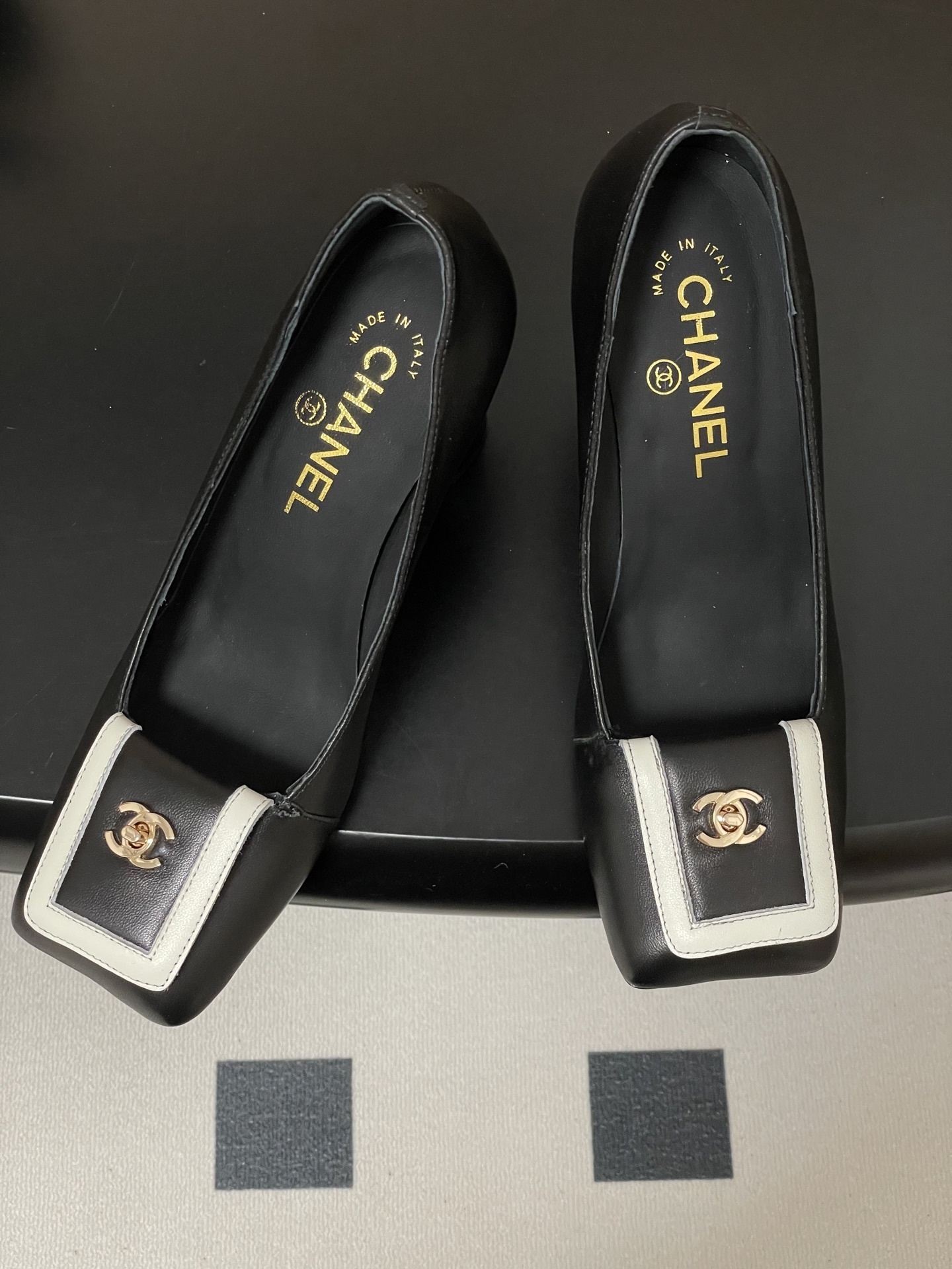 A pair of Chanel black leather block heel pumps with square toes and gold-tone CC logos on white buckle details. - Hình 2