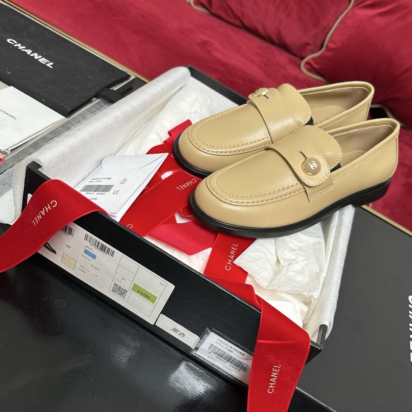 A pair of Chanel beige leather loafers with gold CC logo buttons presented in a luxury black box with red ribbons. - Hình 2