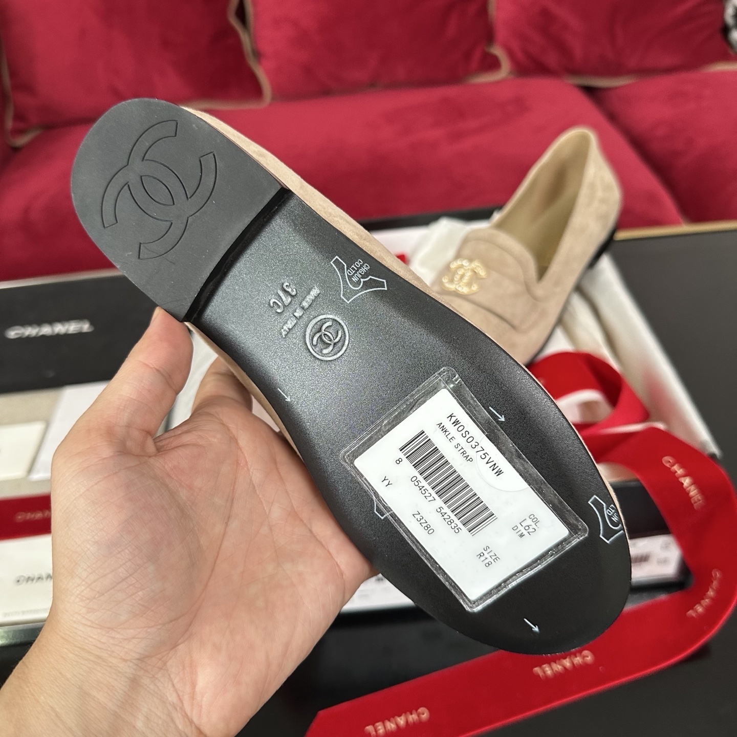 Pair of beige Chanel suede loafers with gold and crystal interlocking CC logos on a black surface with red ribbons. - Hình 9