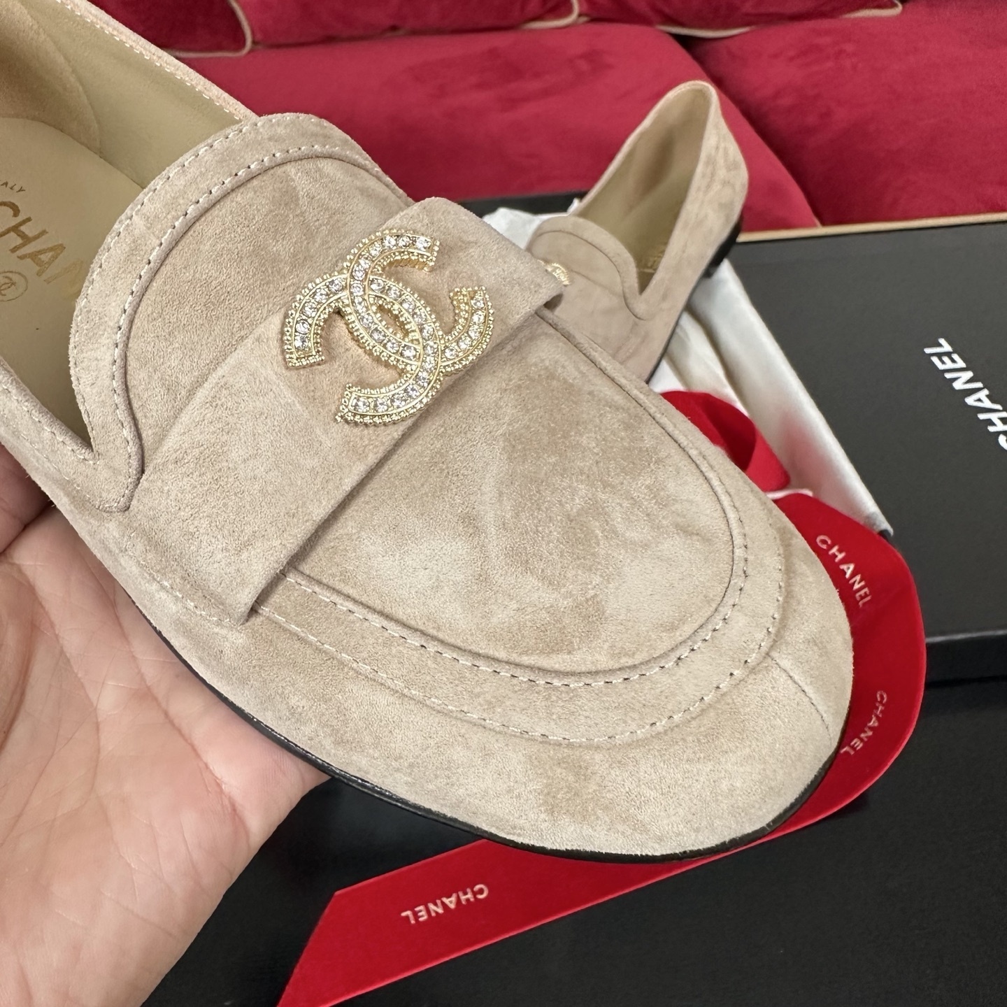 Pair of beige Chanel suede loafers with gold and crystal interlocking CC logos on a black surface with red ribbons. - Hình 6