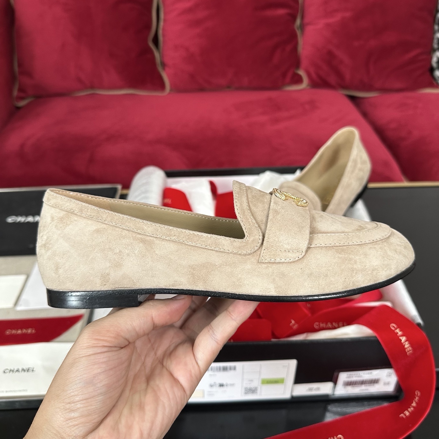 Pair of beige Chanel suede loafers with gold and crystal interlocking CC logos on a black surface with red ribbons. - Hình 5
