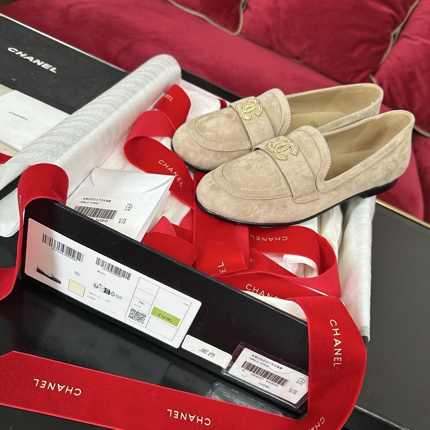 Pair of beige Chanel suede loafers with gold and crystal interlocking CC logos on a black surface with red ribbons. - Hình 2