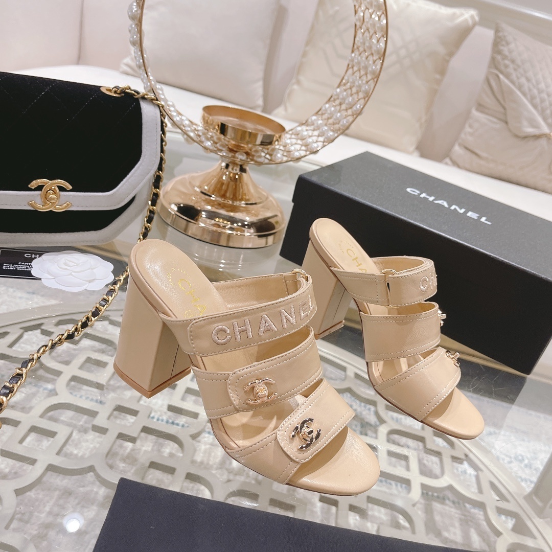 A pair of beige Chanel triple strap block heel mules with gold CC logos and embroidered branding displayed on a glass table. - Hình 3
