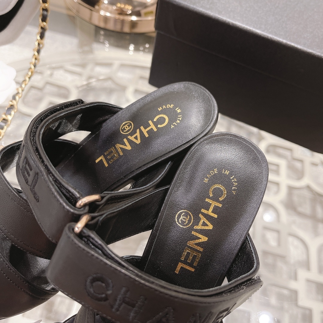 A pair of luxury Chanel black leather triple strap block heel mules with gold CC logos displayed on a glass table - Hình 7