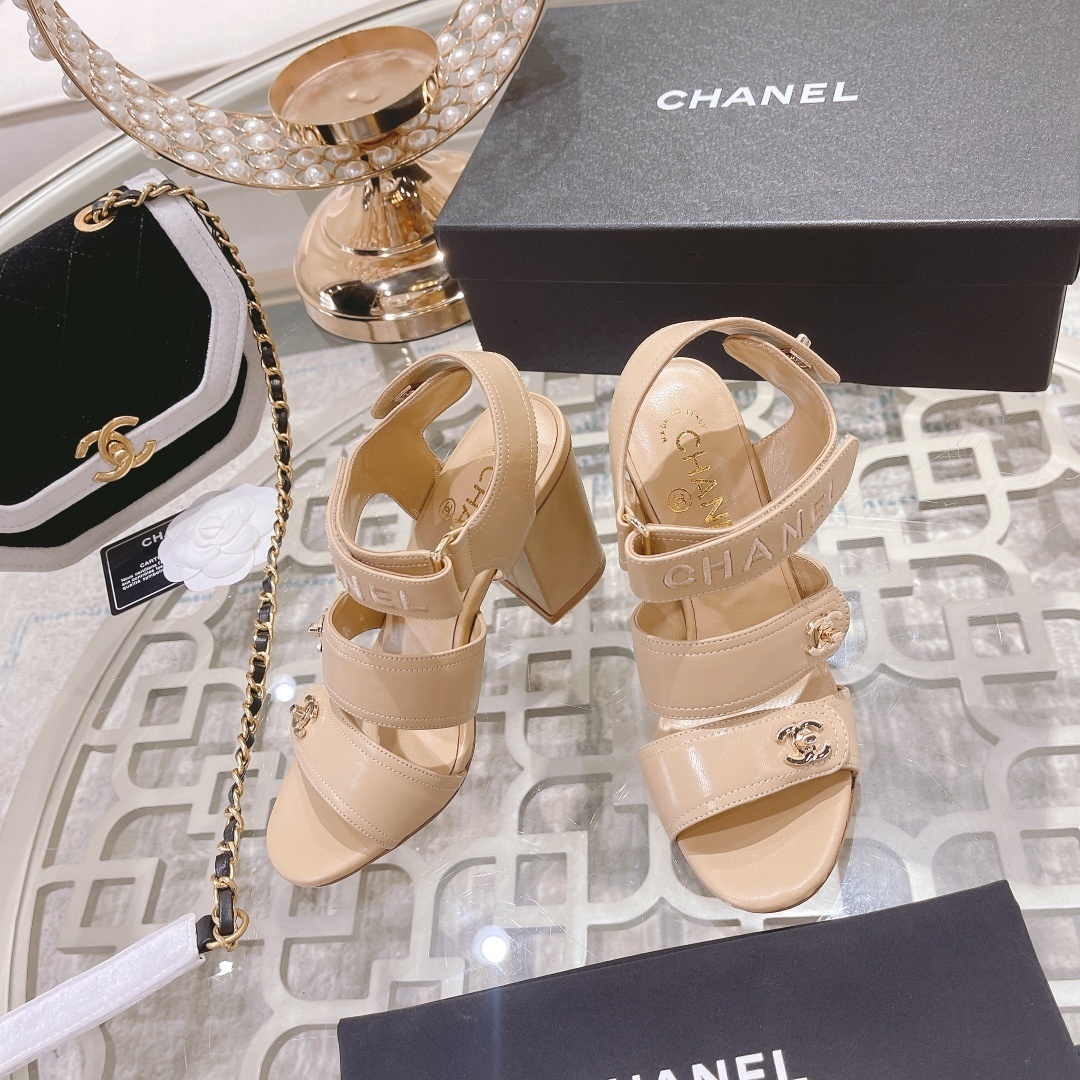 Pair of Chanel beige leather block heel sandals with gold CC logo hardware and branded ankle straps displayed on a glass table. - Hình 2