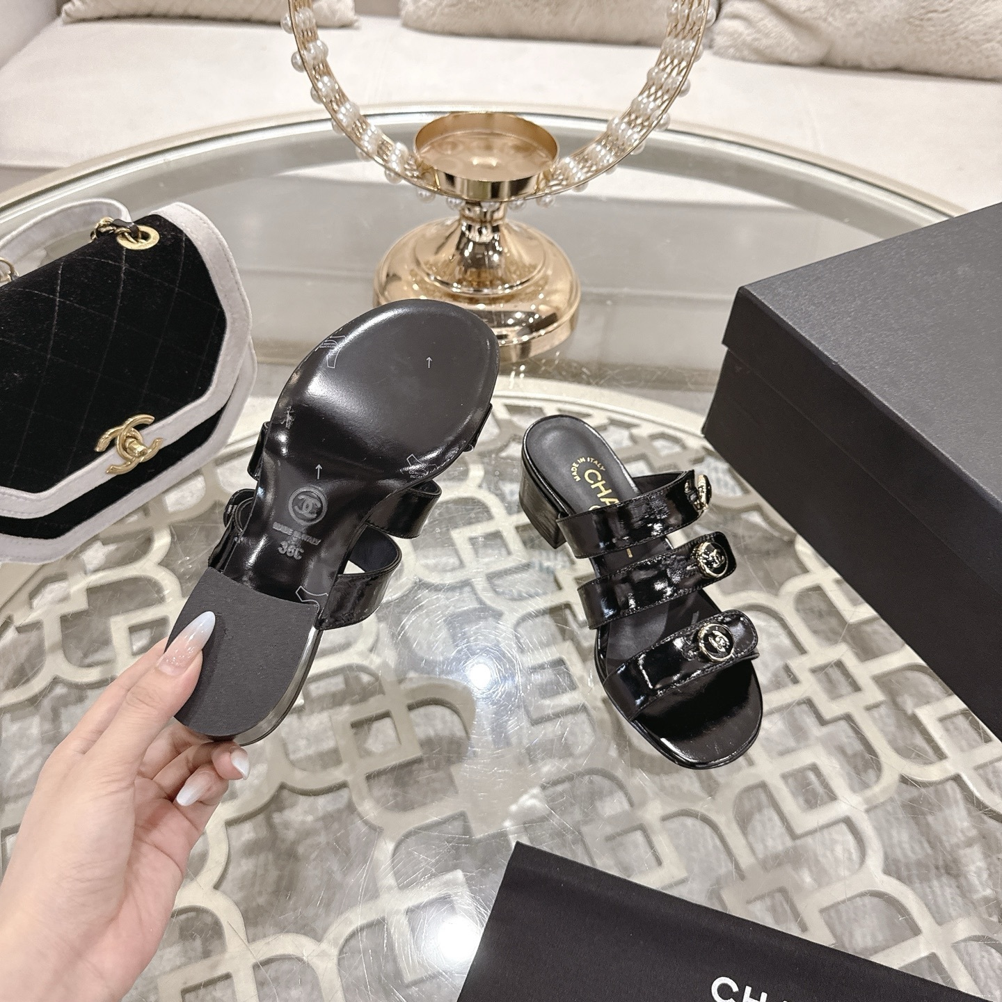 A pair of Chanel black patent leather sandals with three straps and gold CC logo buttons displayed on a glass table. - Hình 9