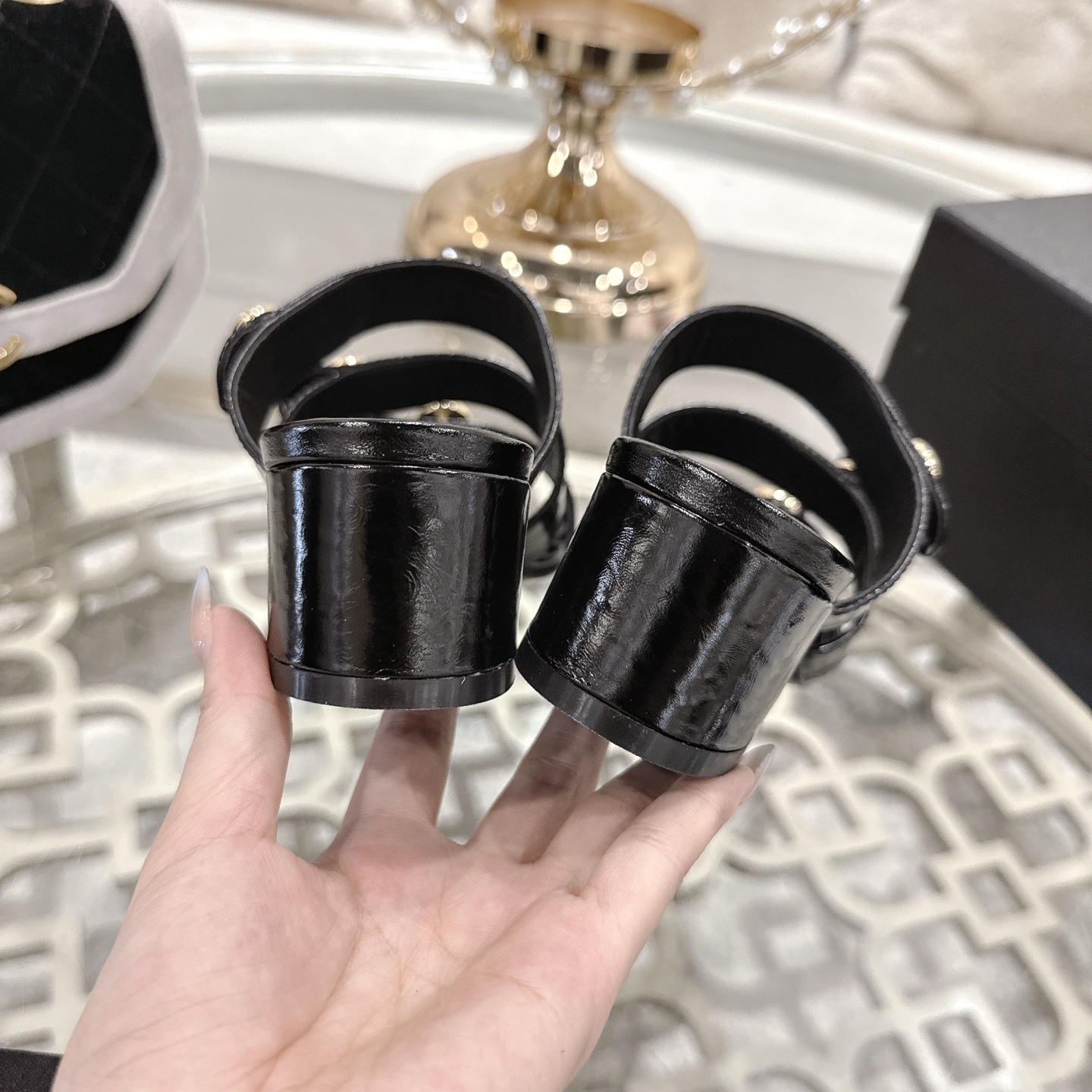 A pair of Chanel black patent leather sandals with three straps and gold CC logo buttons displayed on a glass table. - Hình 8