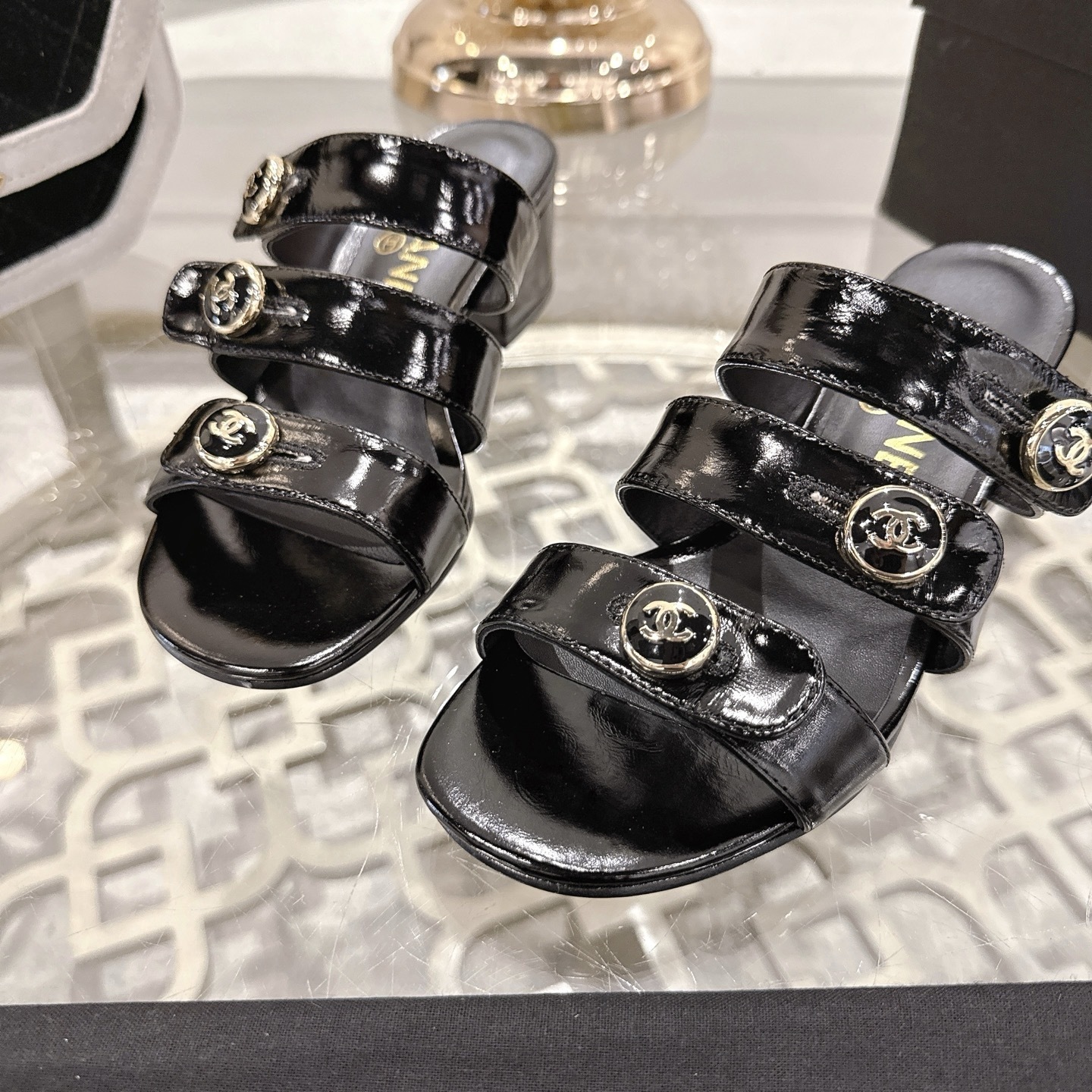 A pair of Chanel black patent leather sandals with three straps and gold CC logo buttons displayed on a glass table. - Hình 7