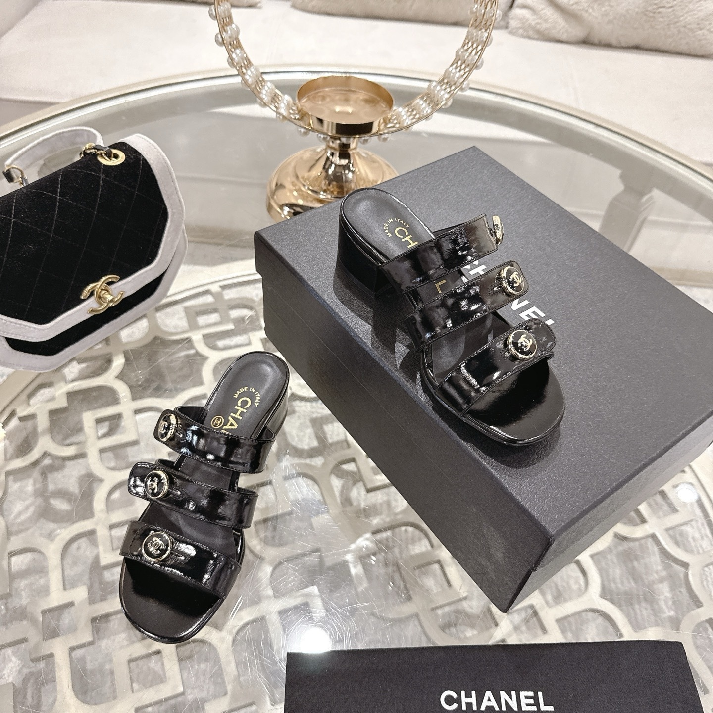 A pair of Chanel black patent leather sandals with three straps and gold CC logo buttons displayed on a glass table. - Hình 6