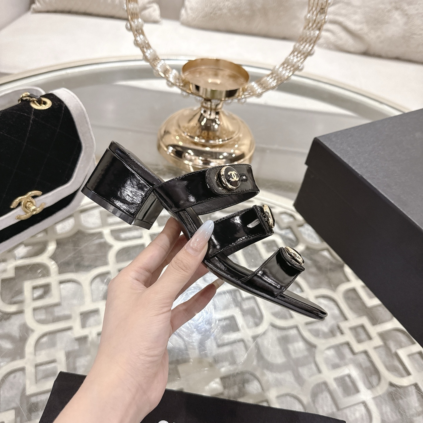 A pair of Chanel black patent leather sandals with three straps and gold CC logo buttons displayed on a glass table. - Hình 5