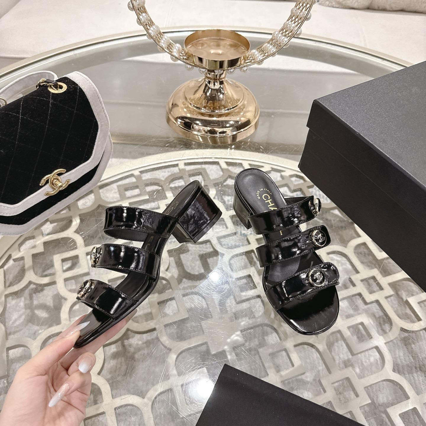 A pair of Chanel black patent leather sandals with three straps and gold CC logo buttons displayed on a glass table. - Hình 4