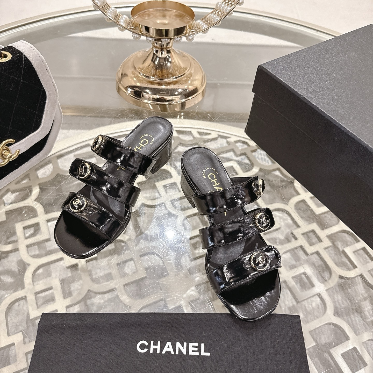 A pair of Chanel black patent leather sandals with three straps and gold CC logo buttons displayed on a glass table. - Hình 2