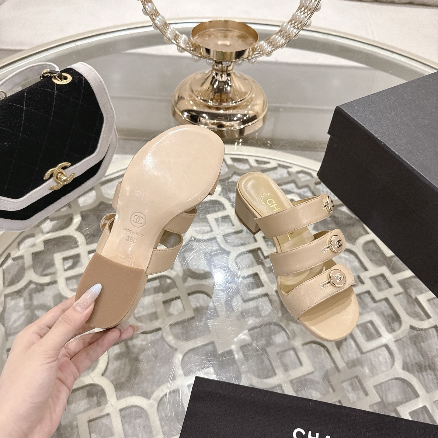 Luxury beige leather sandals with three straps, gold CC logo buttons, and a low block heel displayed on a decorative glass table. - Hình 9