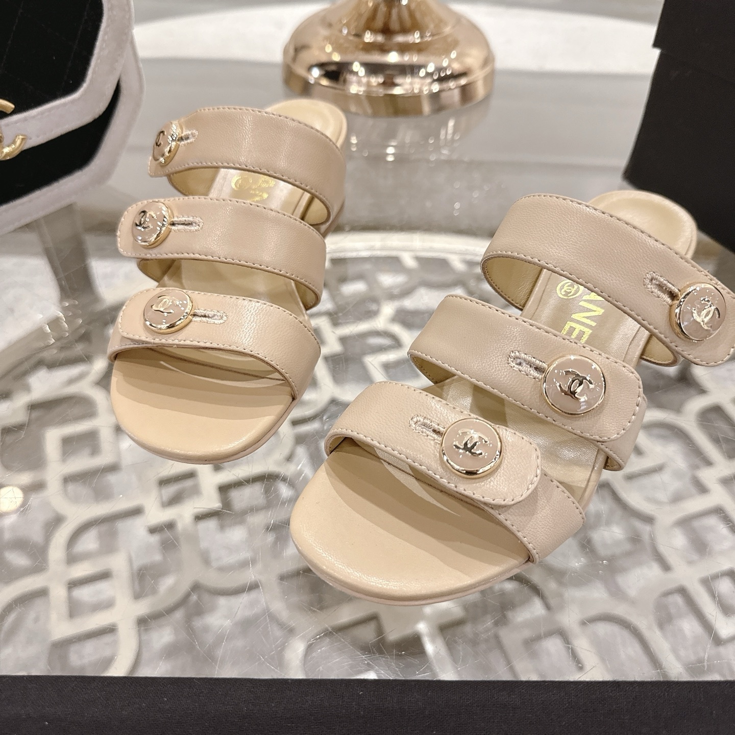 Luxury beige leather sandals with three straps, gold CC logo buttons, and a low block heel displayed on a decorative glass table. - Hình 8