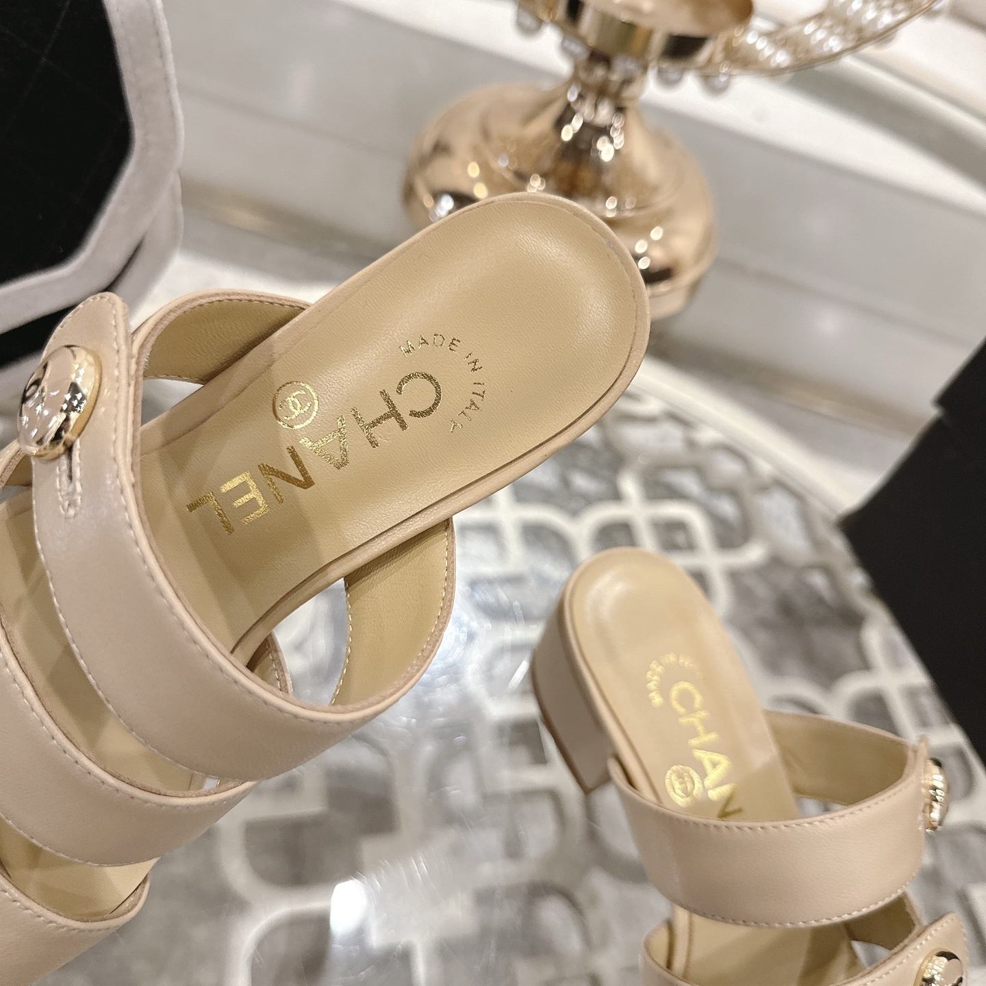 Luxury beige leather sandals with three straps, gold CC logo buttons, and a low block heel displayed on a decorative glass table. - Hình 7