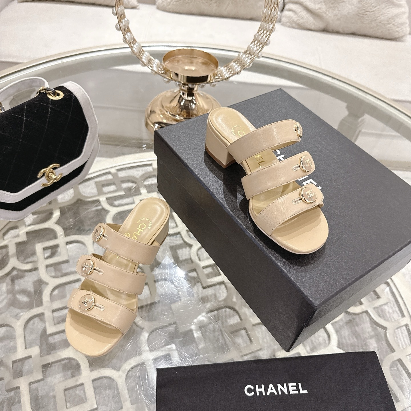 Luxury beige leather sandals with three straps, gold CC logo buttons, and a low block heel displayed on a decorative glass table. - Hình 6