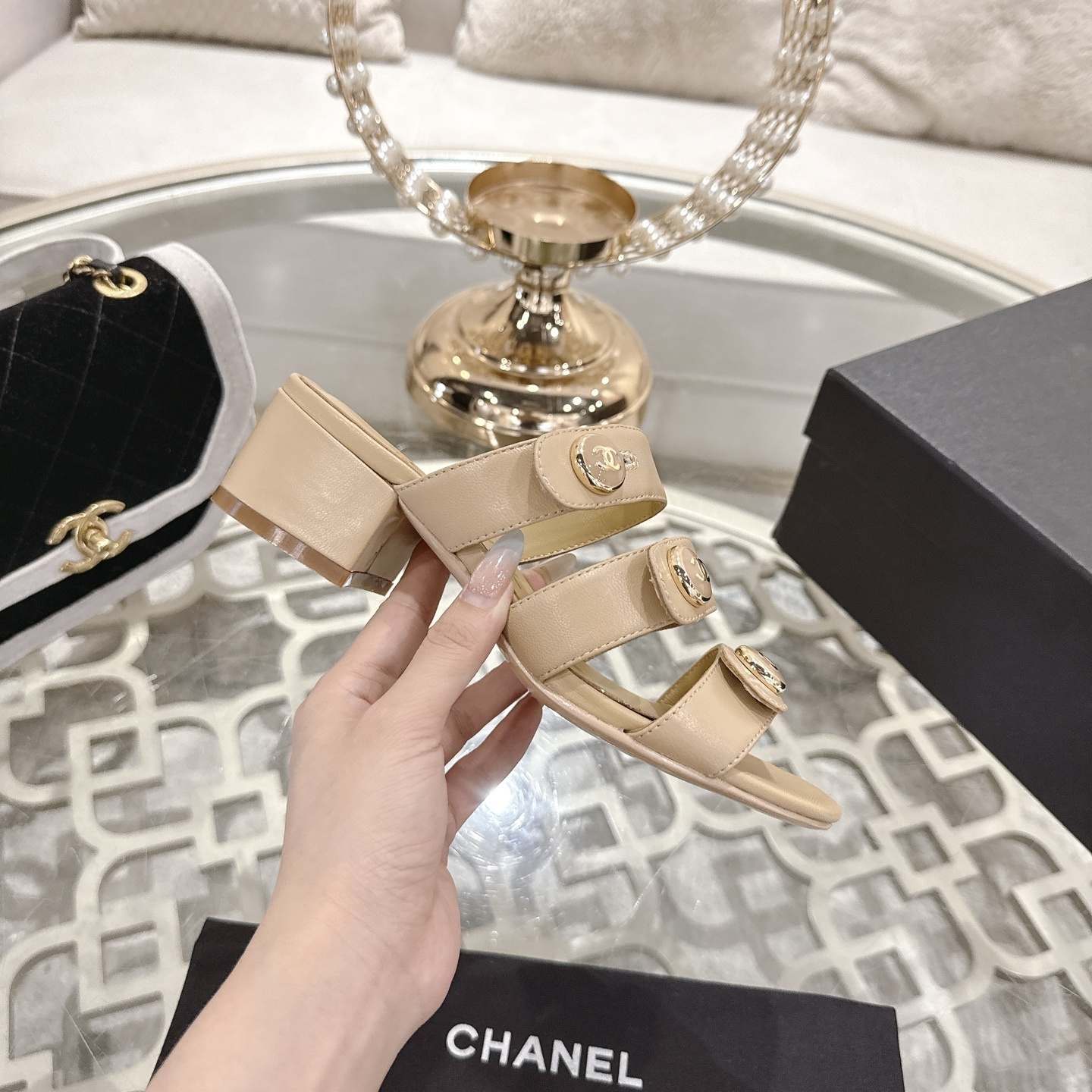 Luxury beige leather sandals with three straps, gold CC logo buttons, and a low block heel displayed on a decorative glass table. - Hình 5