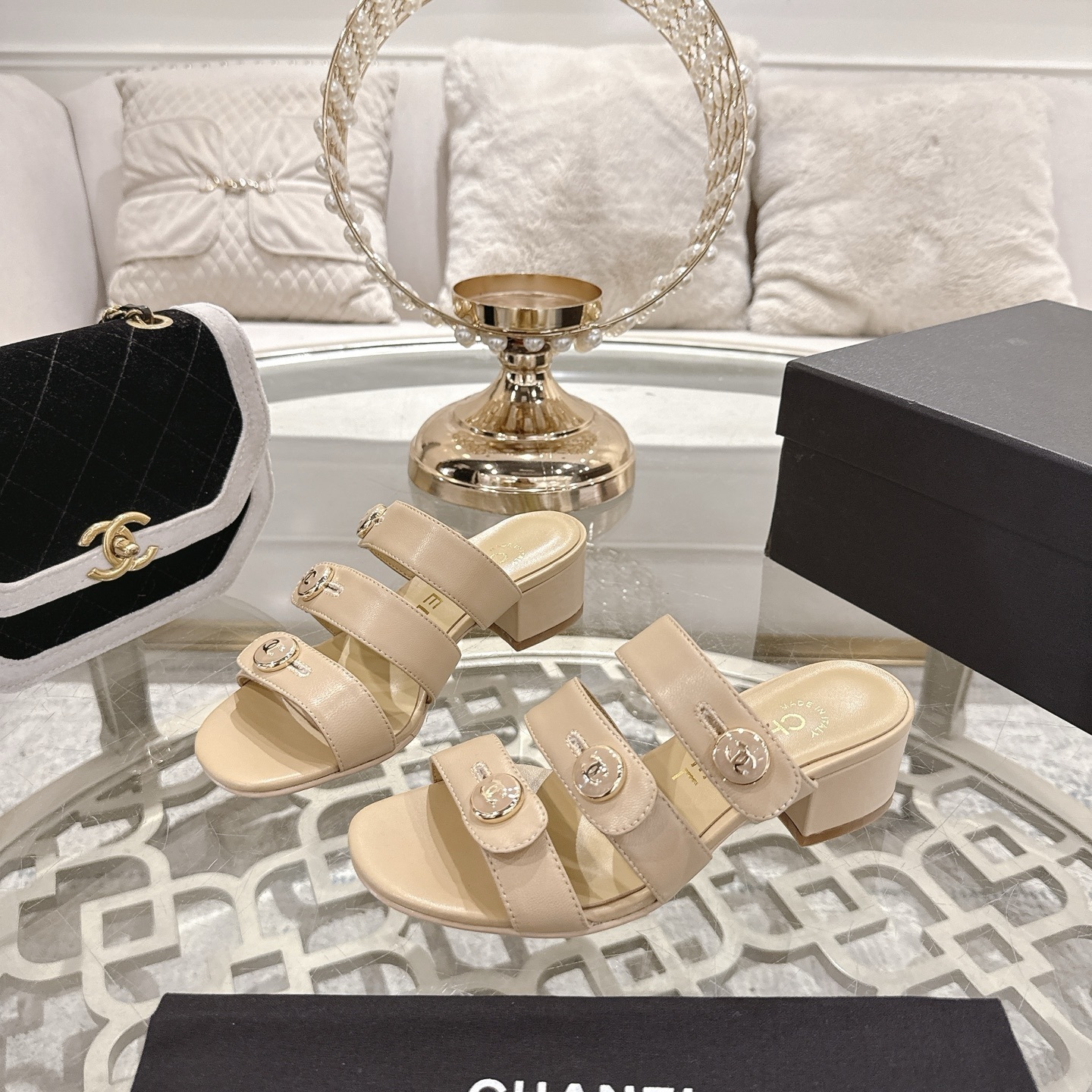 Luxury beige leather sandals with three straps, gold CC logo buttons, and a low block heel displayed on a decorative glass table. - Hình 3