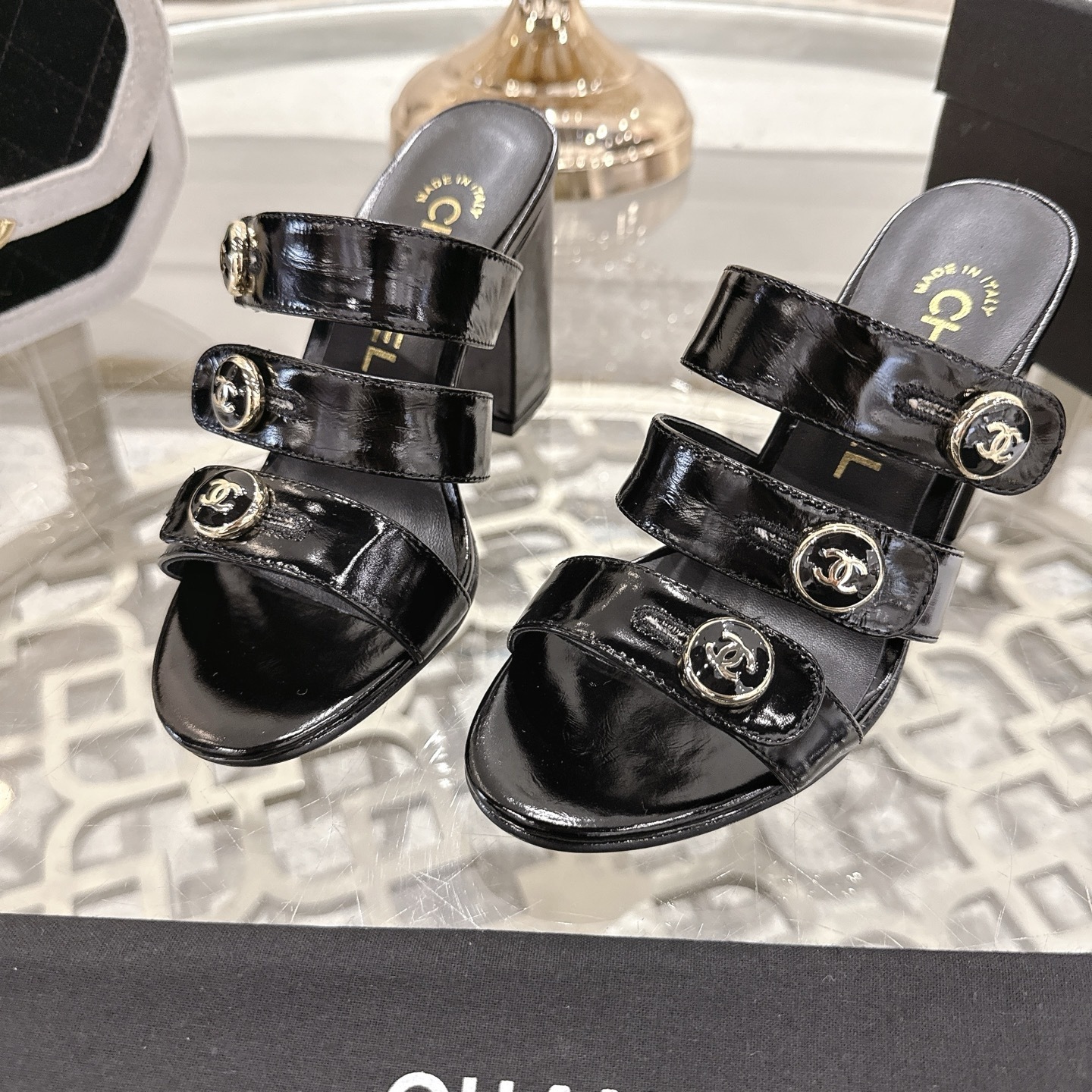 Luxury black patent leather sandals with triple straps and gold CC logo buttons on a designer glass table background. - Hình 7