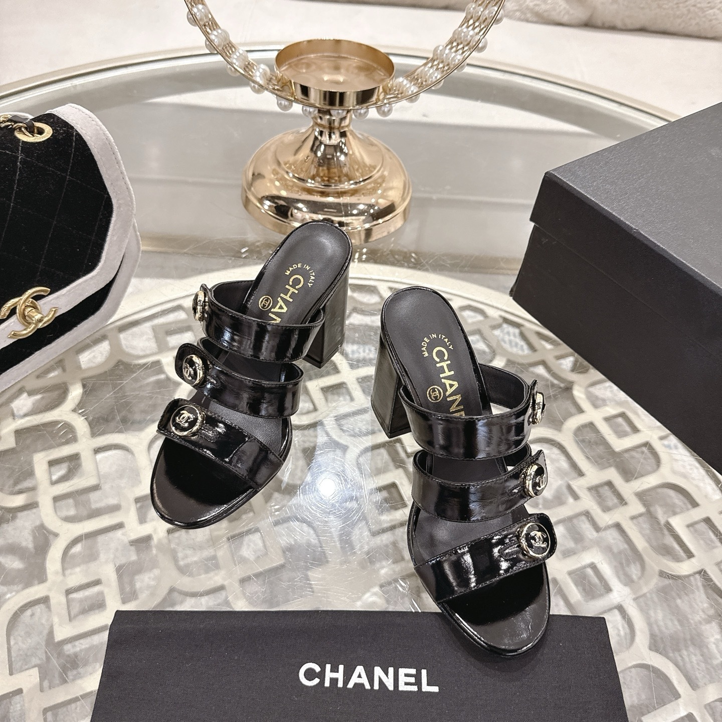 Luxury black patent leather sandals with triple straps and gold CC logo buttons on a designer glass table background. - Hình 2