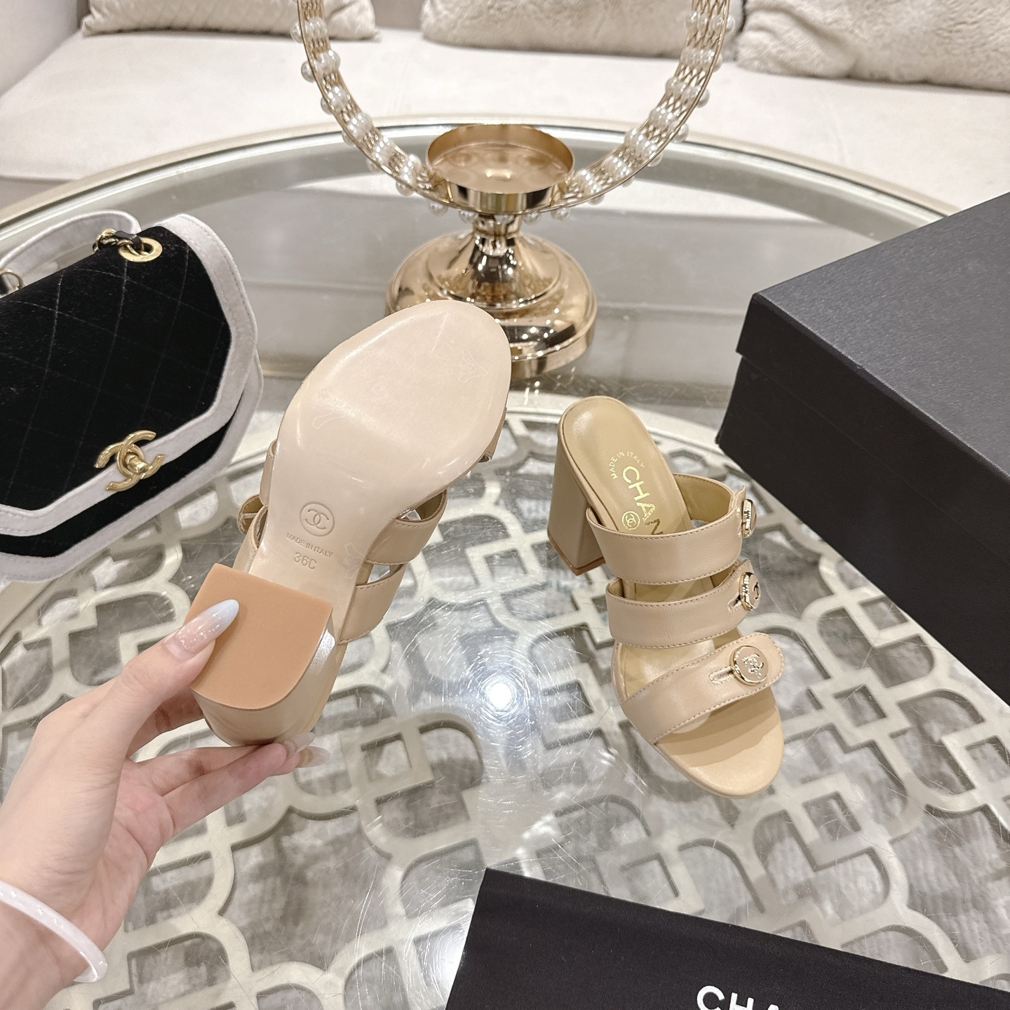 A pair of Chanel beige leather triple strap mules with gold CC logo buttons and block heels displayed on a glass table. - Hình 9