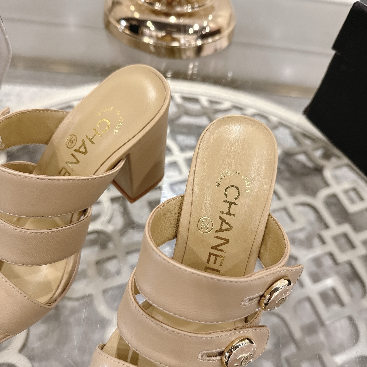A pair of Chanel beige leather triple strap mules with gold CC logo buttons and block heels displayed on a glass table. - Hình 7