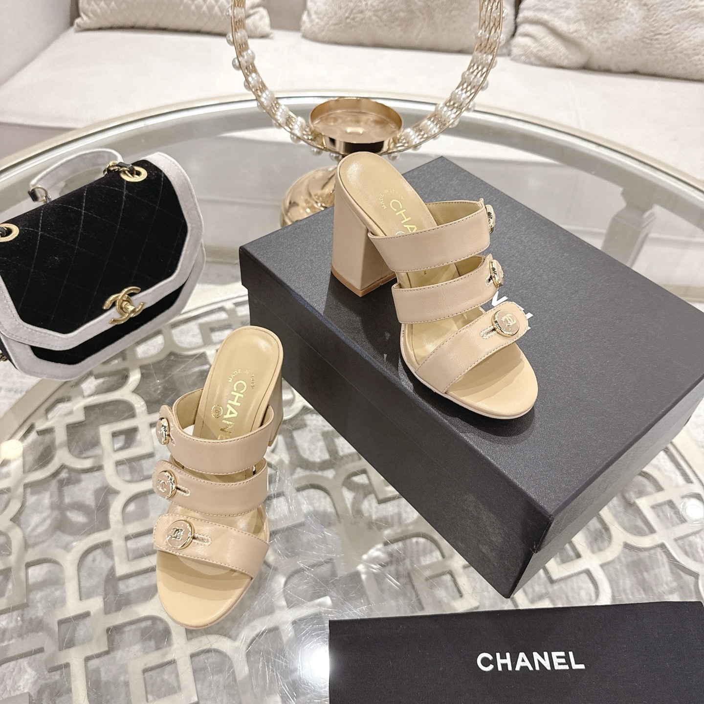 A pair of Chanel beige leather triple strap mules with gold CC logo buttons and block heels displayed on a glass table. - Hình 6