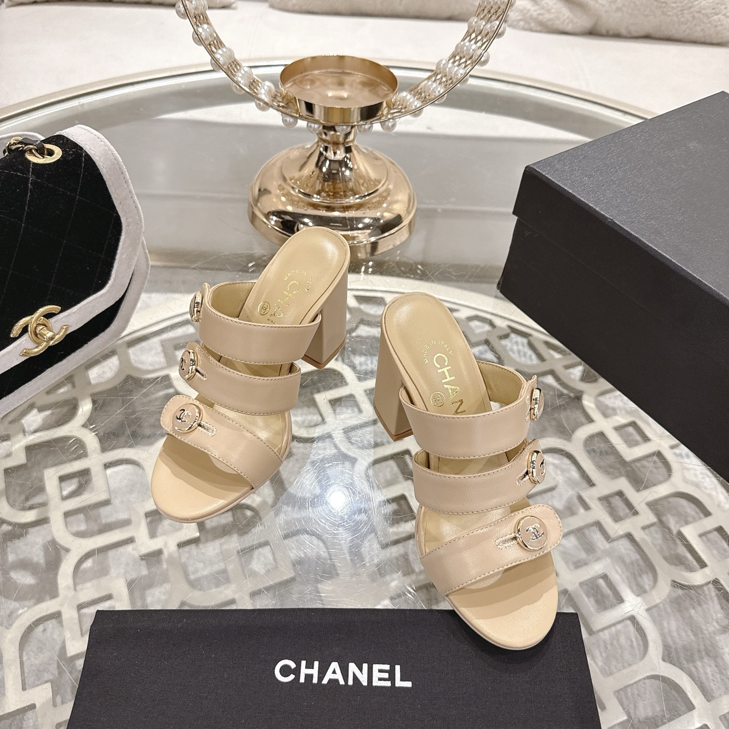 A pair of Chanel beige leather triple strap mules with gold CC logo buttons and block heels displayed on a glass table. - Hình 2