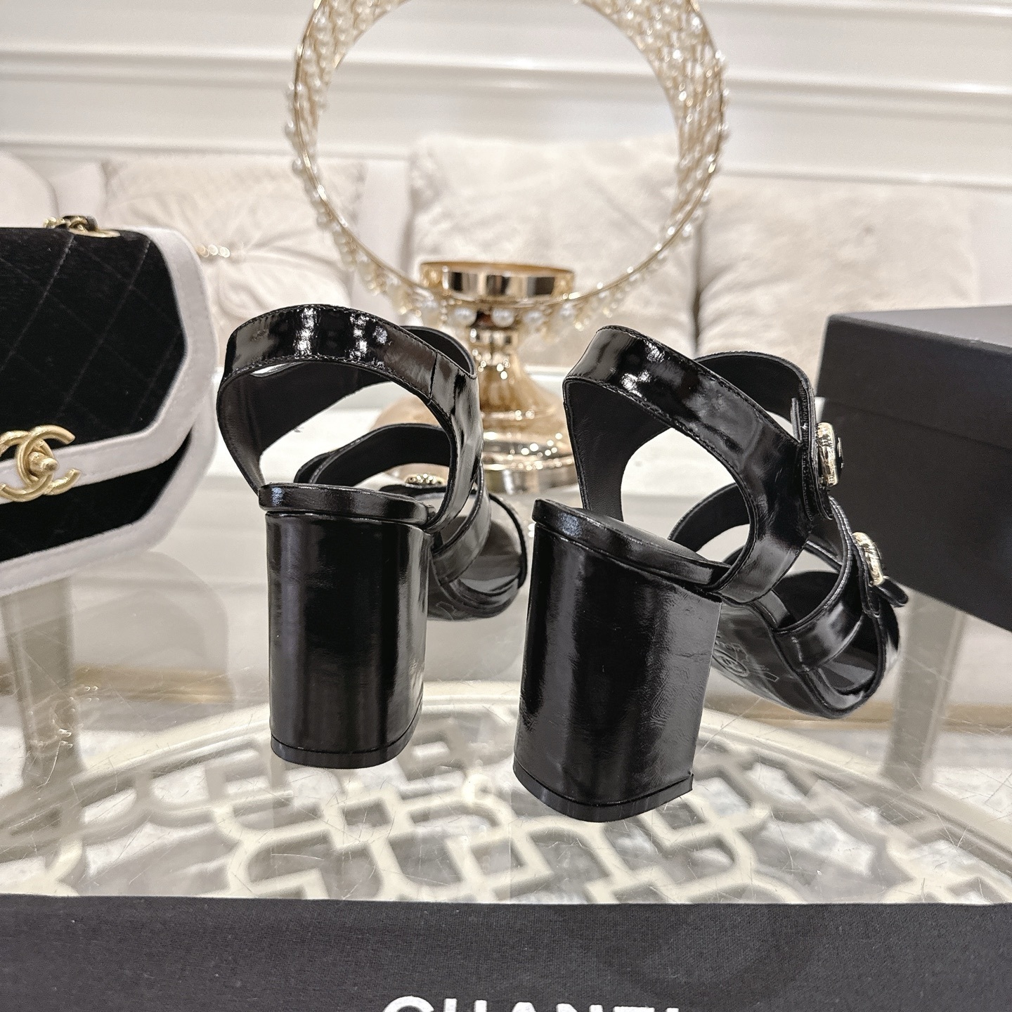 A pair of black patent leather Chanel block heel sandals with gold CC logo buttons displayed on a glass table - Hình 8