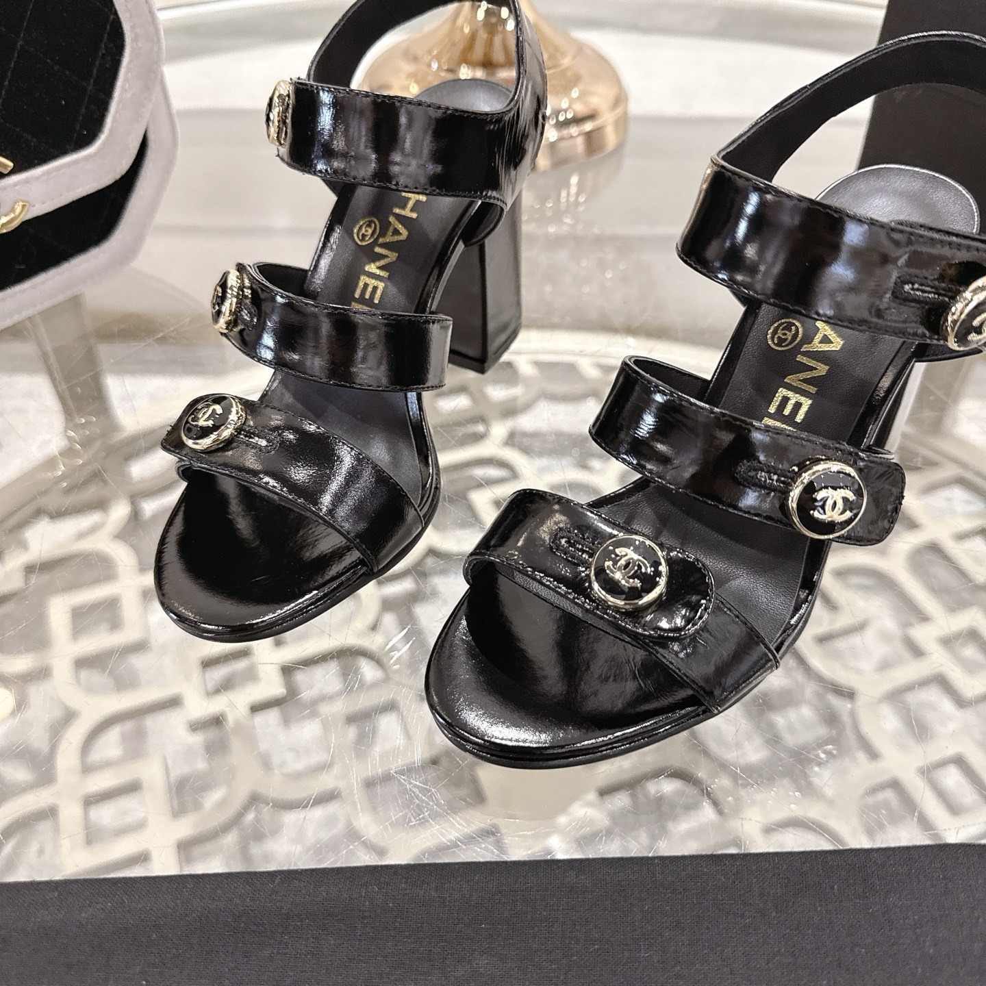 A pair of black patent leather Chanel block heel sandals with gold CC logo buttons displayed on a glass table - Hình 7