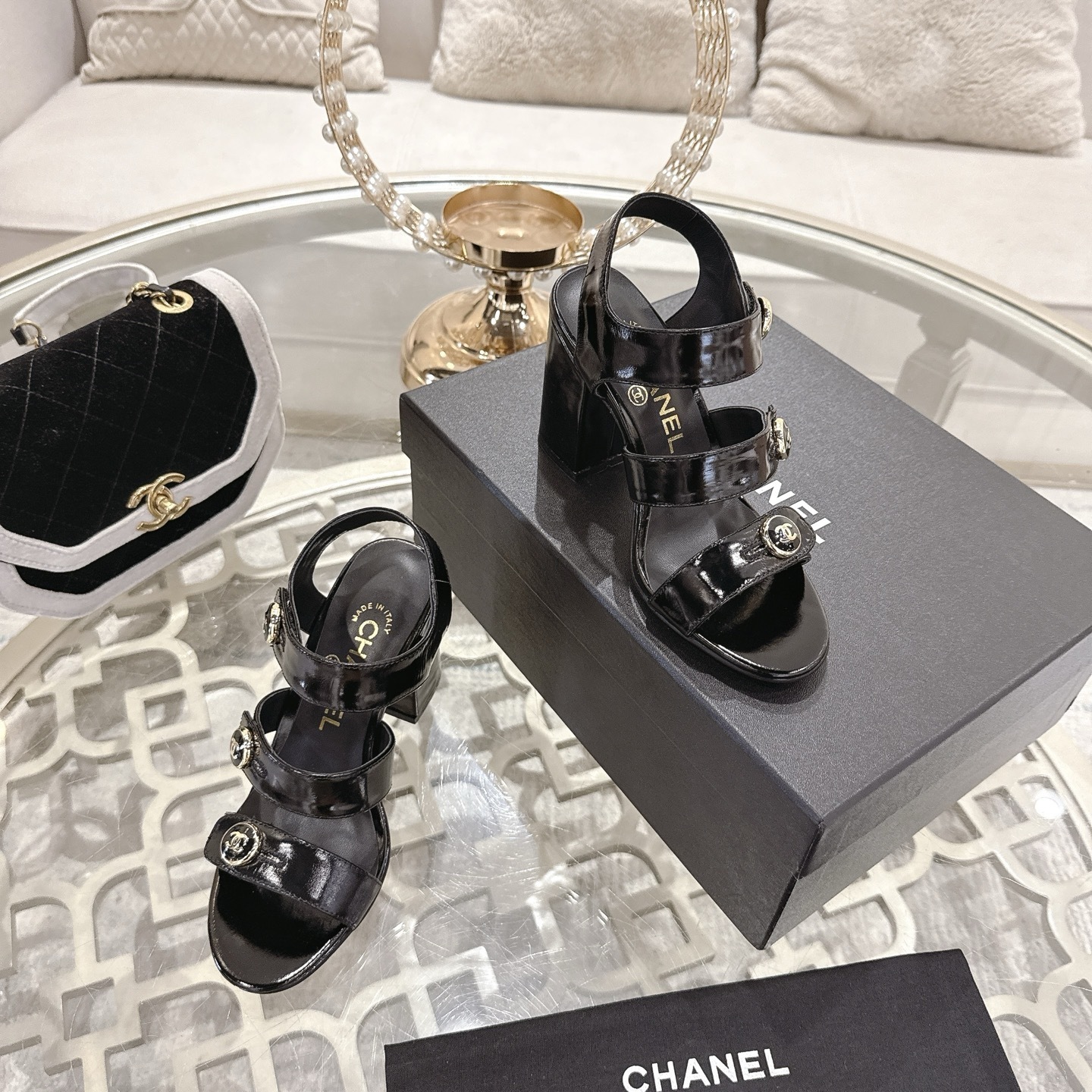 A pair of black patent leather Chanel block heel sandals with gold CC logo buttons displayed on a glass table - Hình 6
