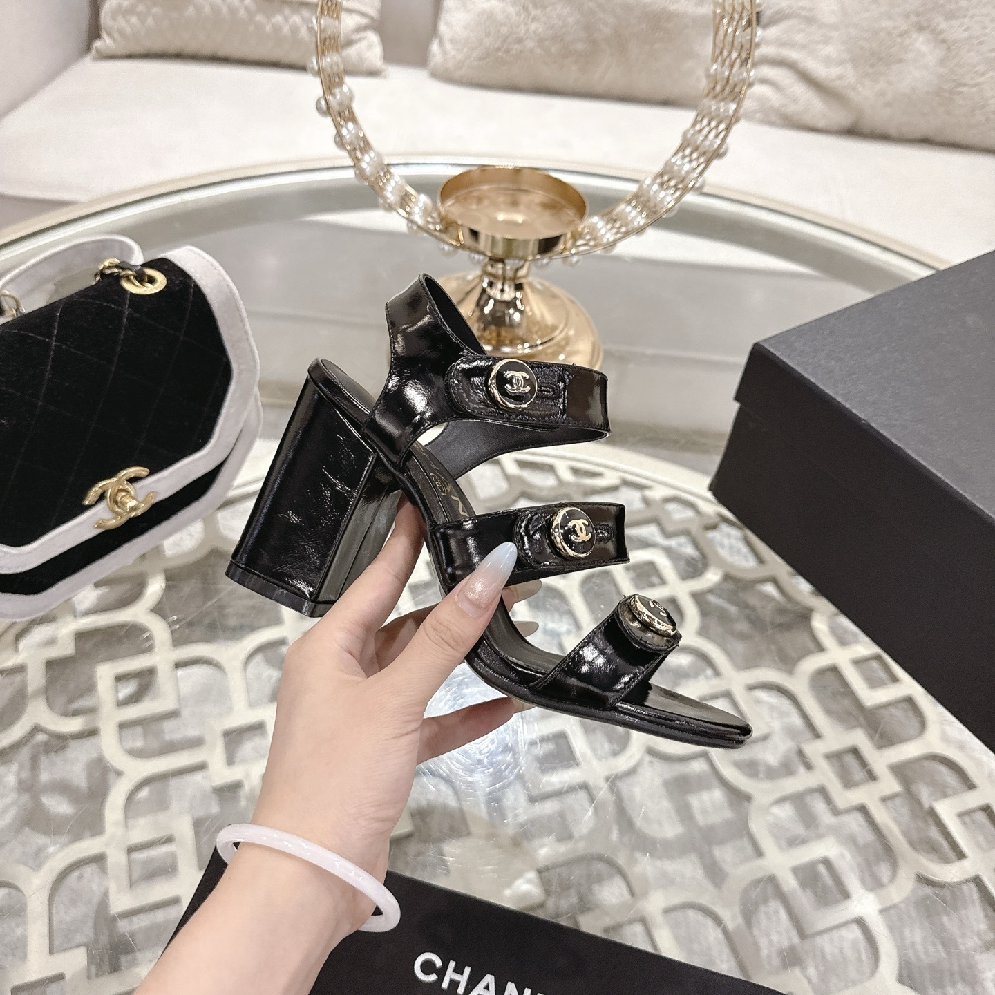 A pair of black patent leather Chanel block heel sandals with gold CC logo buttons displayed on a glass table - Hình 5