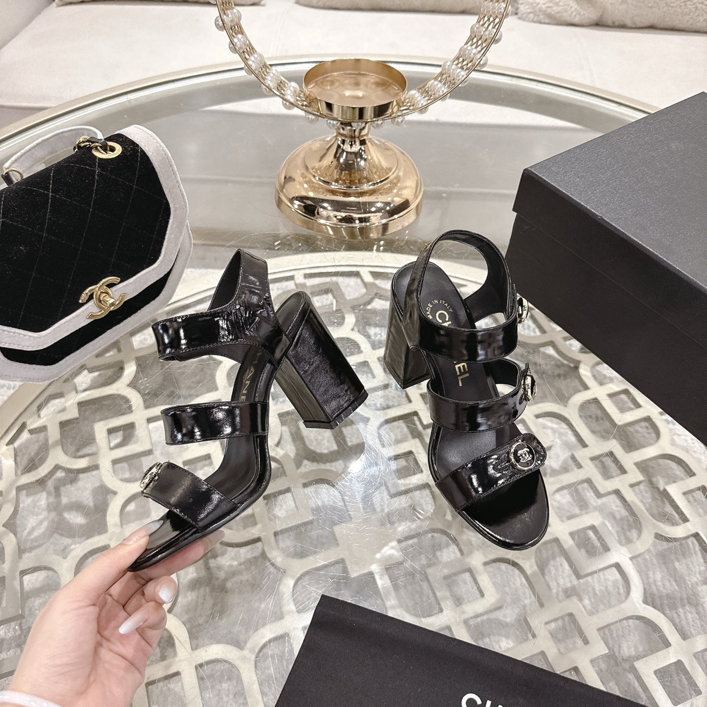 A pair of black patent leather Chanel block heel sandals with gold CC logo buttons displayed on a glass table - Hình 4