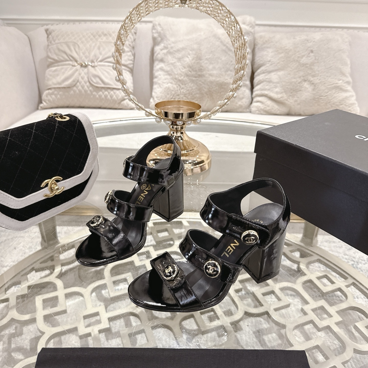 A pair of black patent leather Chanel block heel sandals with gold CC logo buttons displayed on a glass table - Hình 3