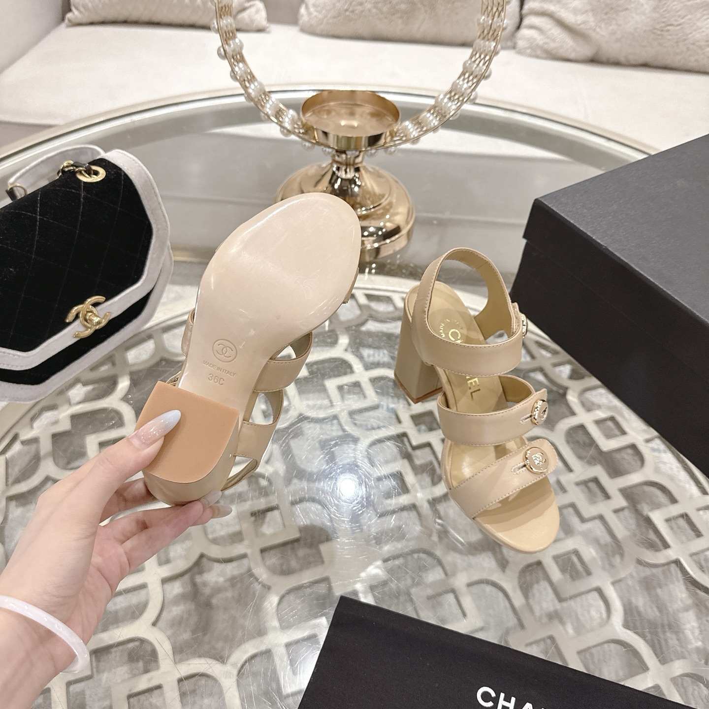 A pair of luxury beige Chanel sandals with three straps and gold CC logo buttons displayed on a glass table. - Hình 9