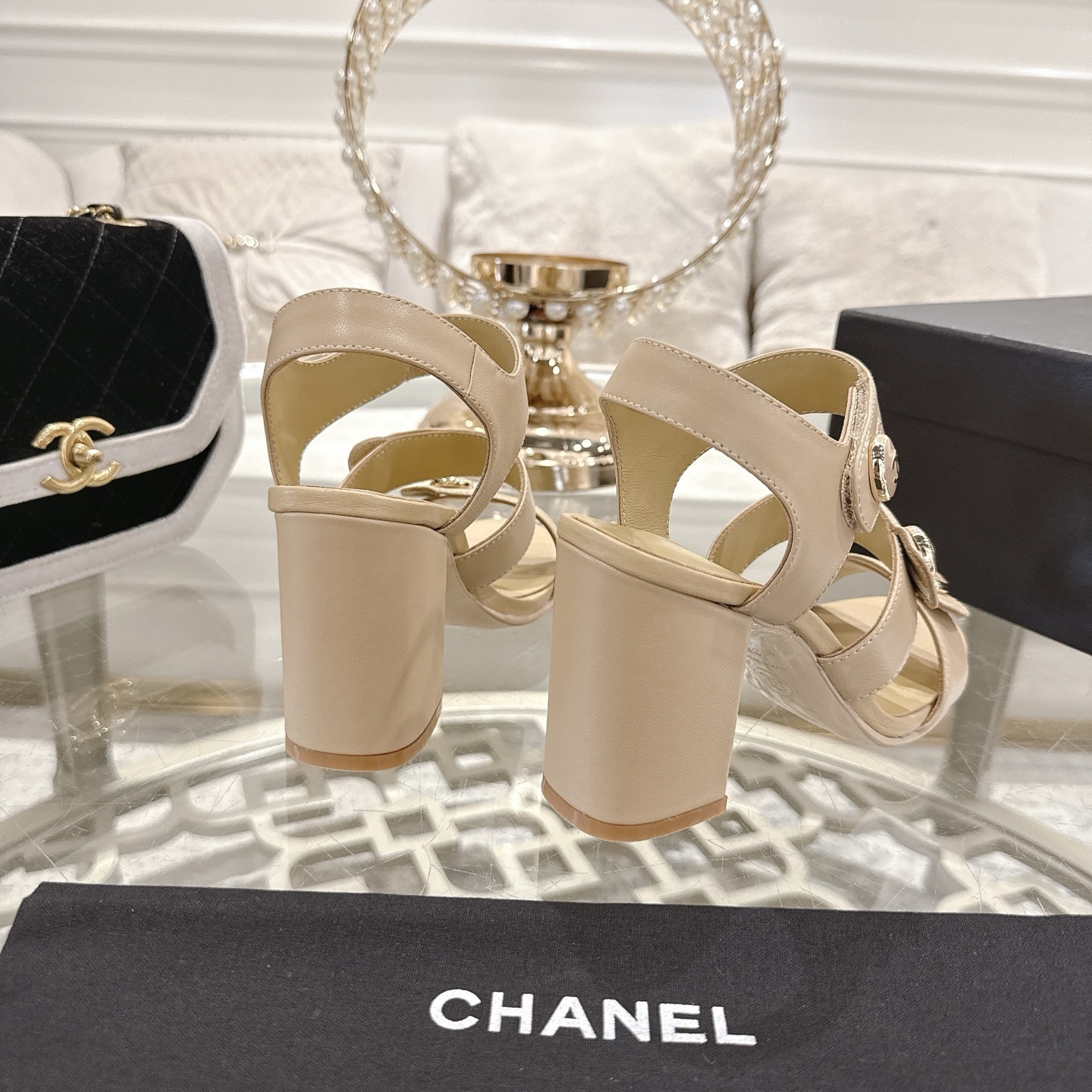 A pair of luxury beige Chanel sandals with three straps and gold CC logo buttons displayed on a glass table. - Hình 8