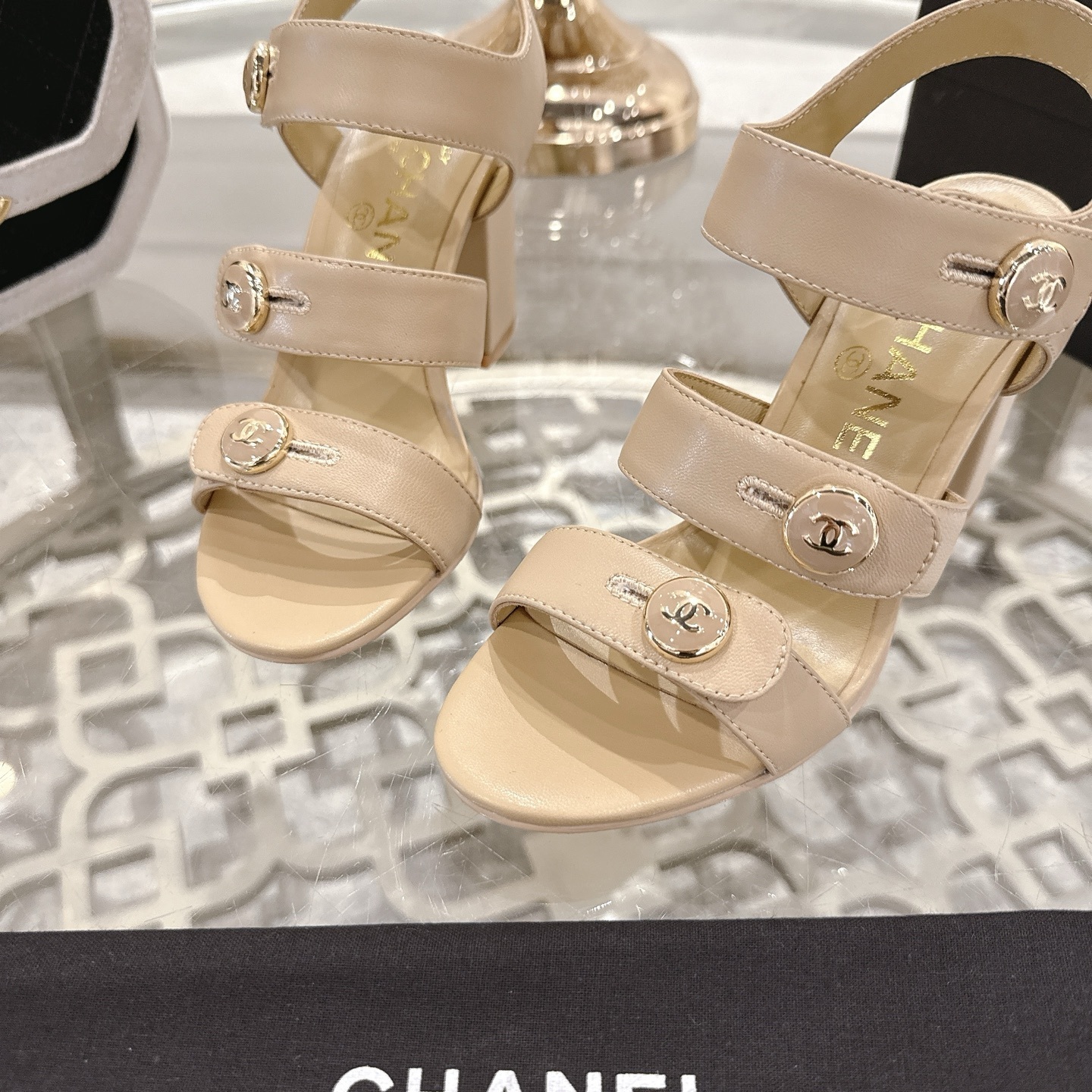 A pair of luxury beige Chanel sandals with three straps and gold CC logo buttons displayed on a glass table. - Hình 7