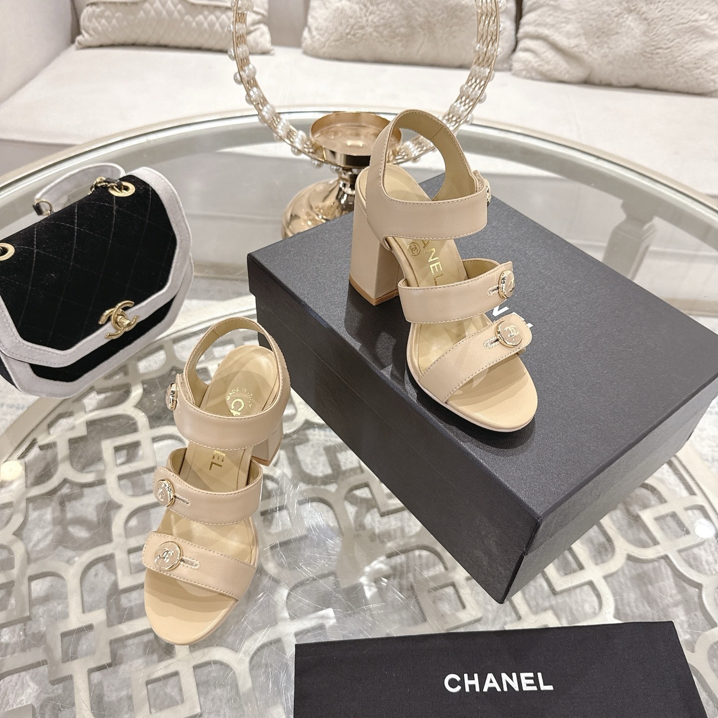A pair of luxury beige Chanel sandals with three straps and gold CC logo buttons displayed on a glass table. - Hình 6