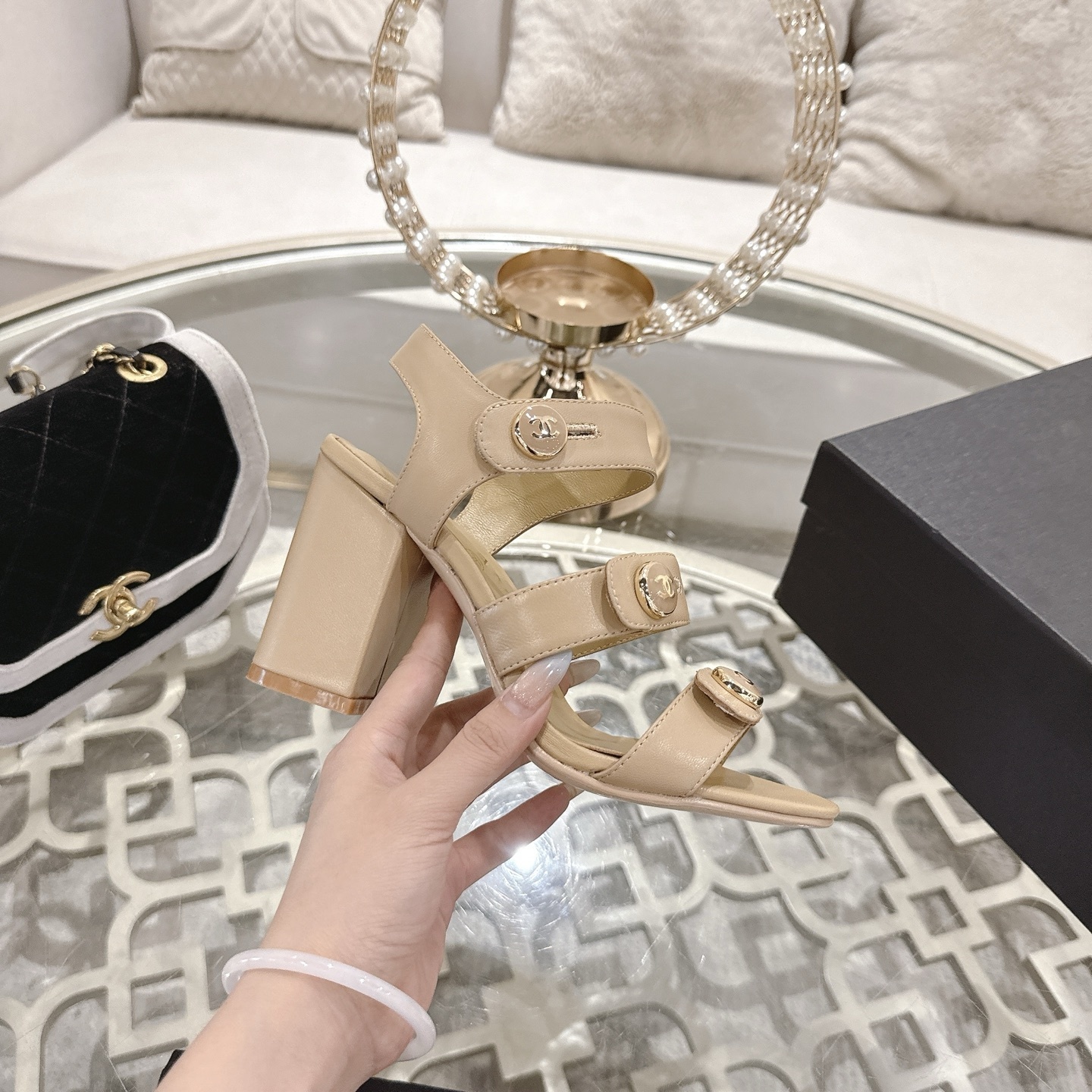 A pair of luxury beige Chanel sandals with three straps and gold CC logo buttons displayed on a glass table. - Hình 5