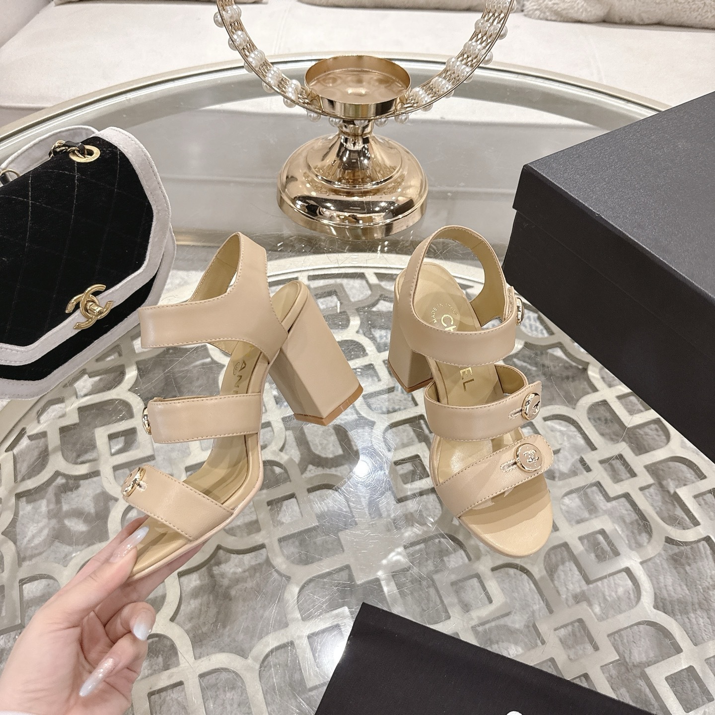 A pair of luxury beige Chanel sandals with three straps and gold CC logo buttons displayed on a glass table. - Hình 4