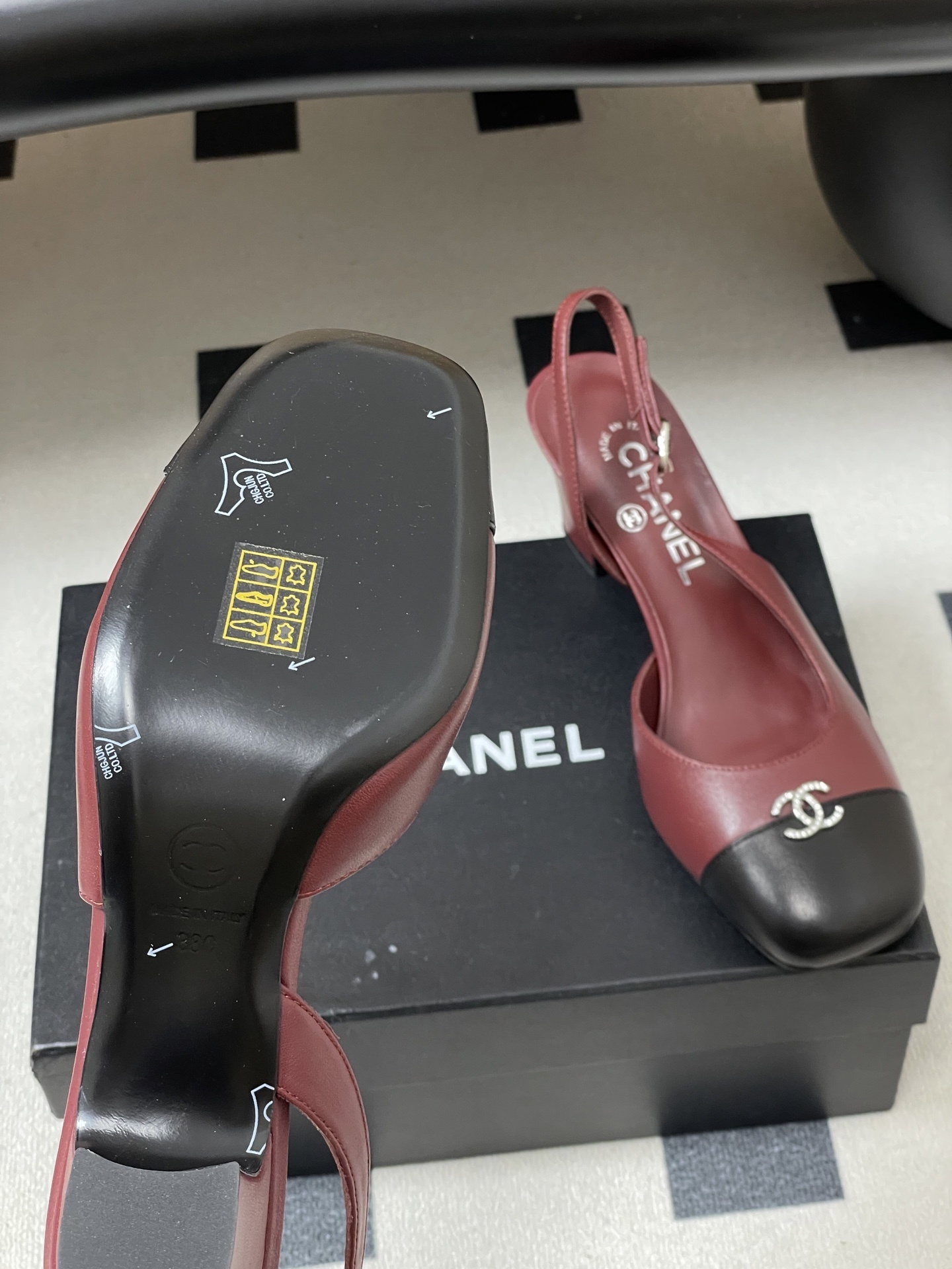A pair of Chanel burgundy leather slingback heels with black cap toes and crystal-embellished CC logos on a black surface. - Hình 8
